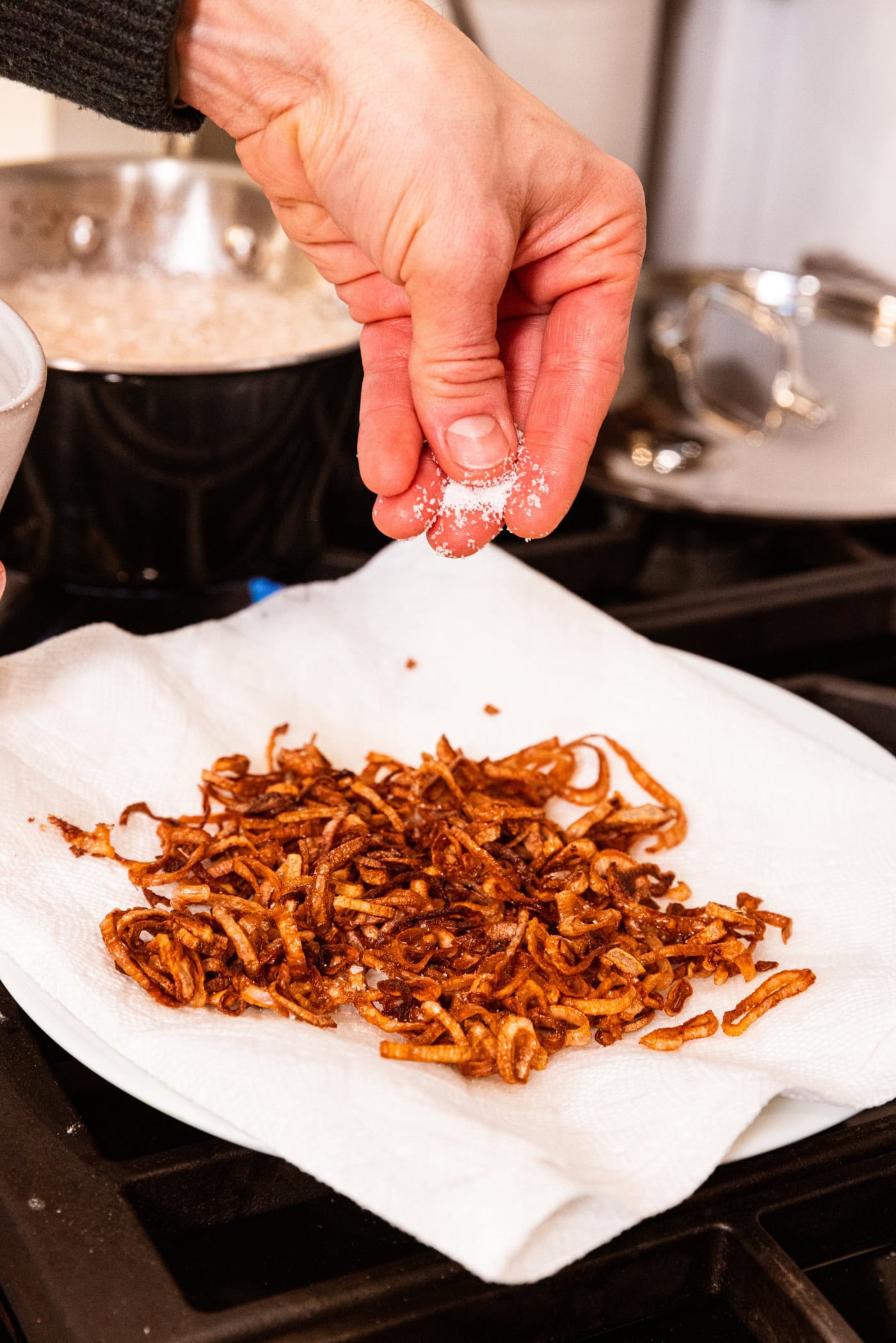 Hand sprinkling salt on top of crispy fried scallions resting on top of a paper towel on a plate.