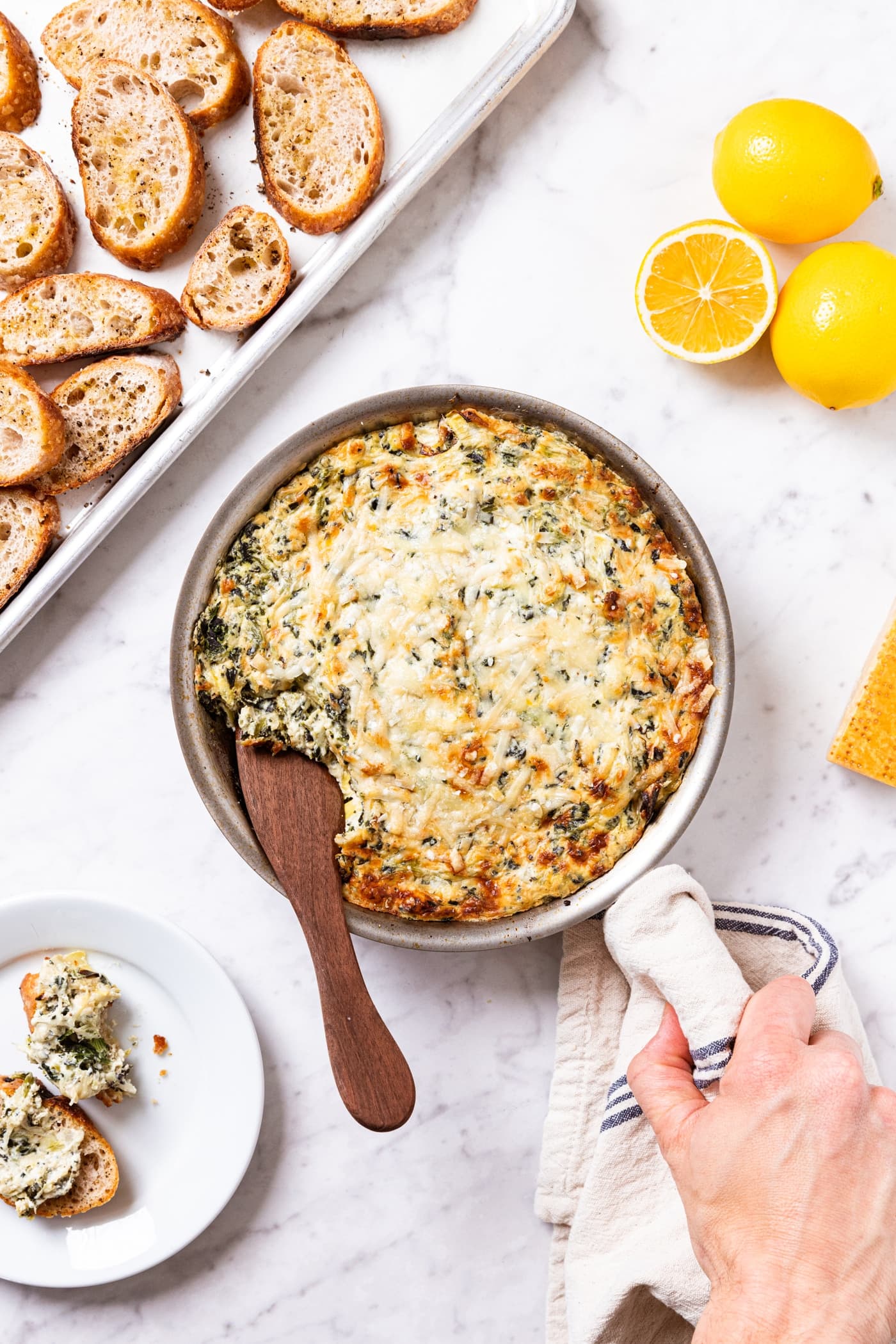 Hand holding handle of metal skillet filled with baked spinach artichoke dip sitting on white marble surface with wooden spoon taking a scoop out along with lemons and crostini to the sides.