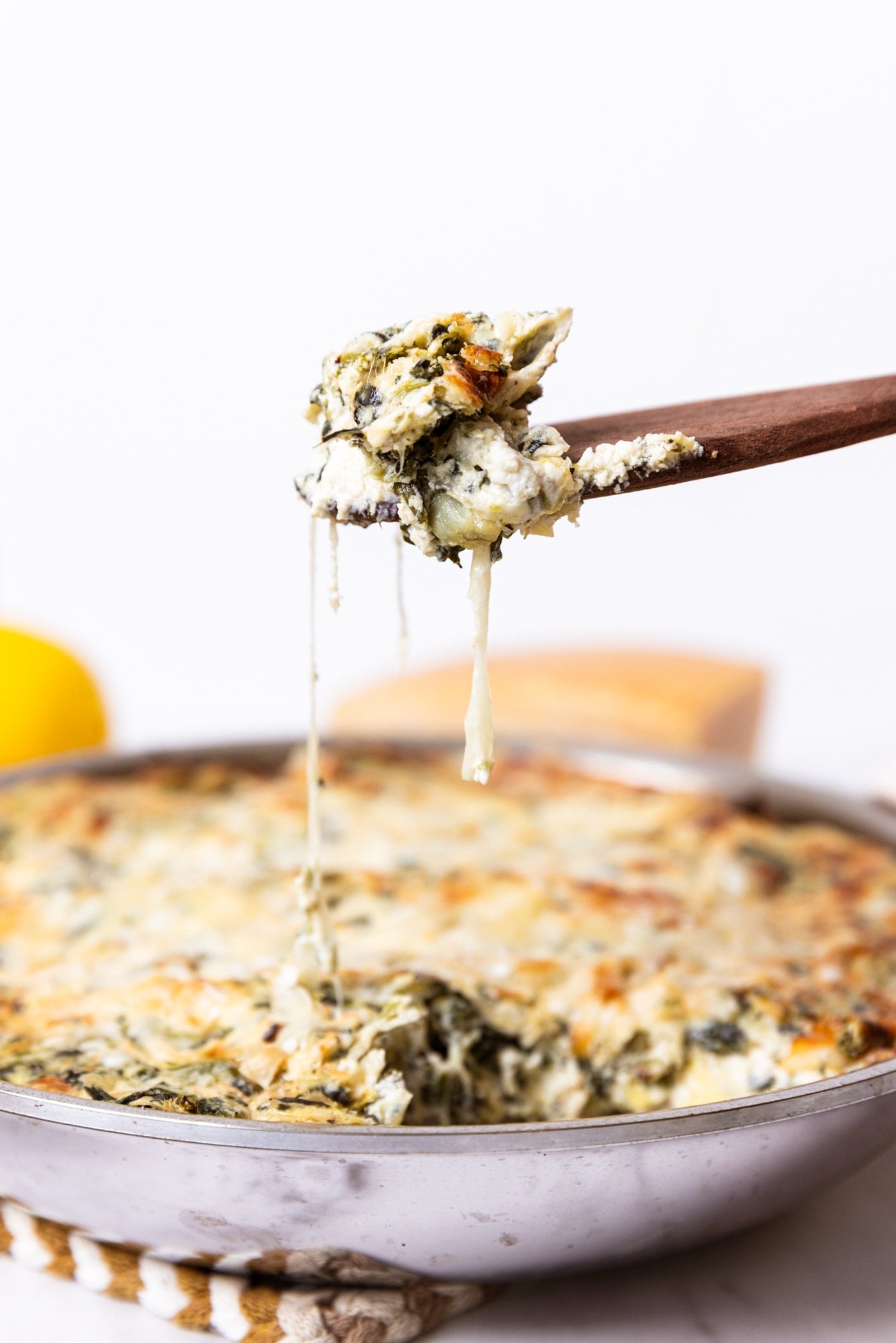 Wooden spatula holding up scoop of spinach artichoke dip with strings of cheese hanging down to rest of dip in metal skillet below.