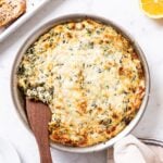 Metal skillet filled with baked spinach artichoke dip with brown top sitting on white marble surface with lemons, Parmesan, and crostini around.