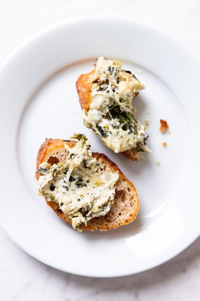 White plate with two pieces of crostini topped with a serving of baked spinach artichoke dip on top.