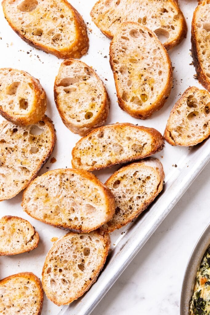 Baked crostini sitting on baking sheet after coming out of the oven showing their crispy texture and edges sitting on white marble surface.
