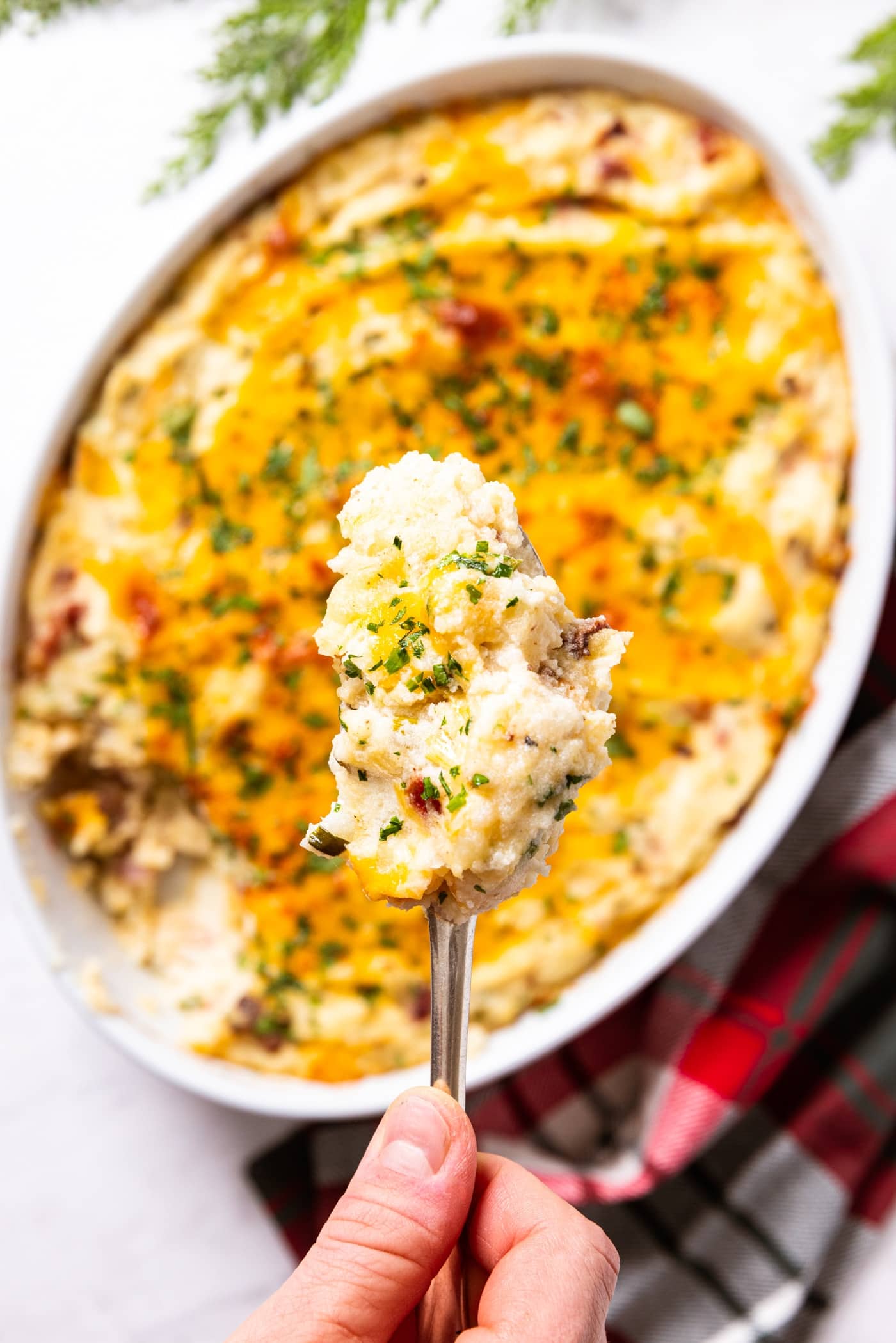 Hand holding spoon filled with serving of potato casserole with white baking dish filled with rest of casserole below sitting on white marble surface.