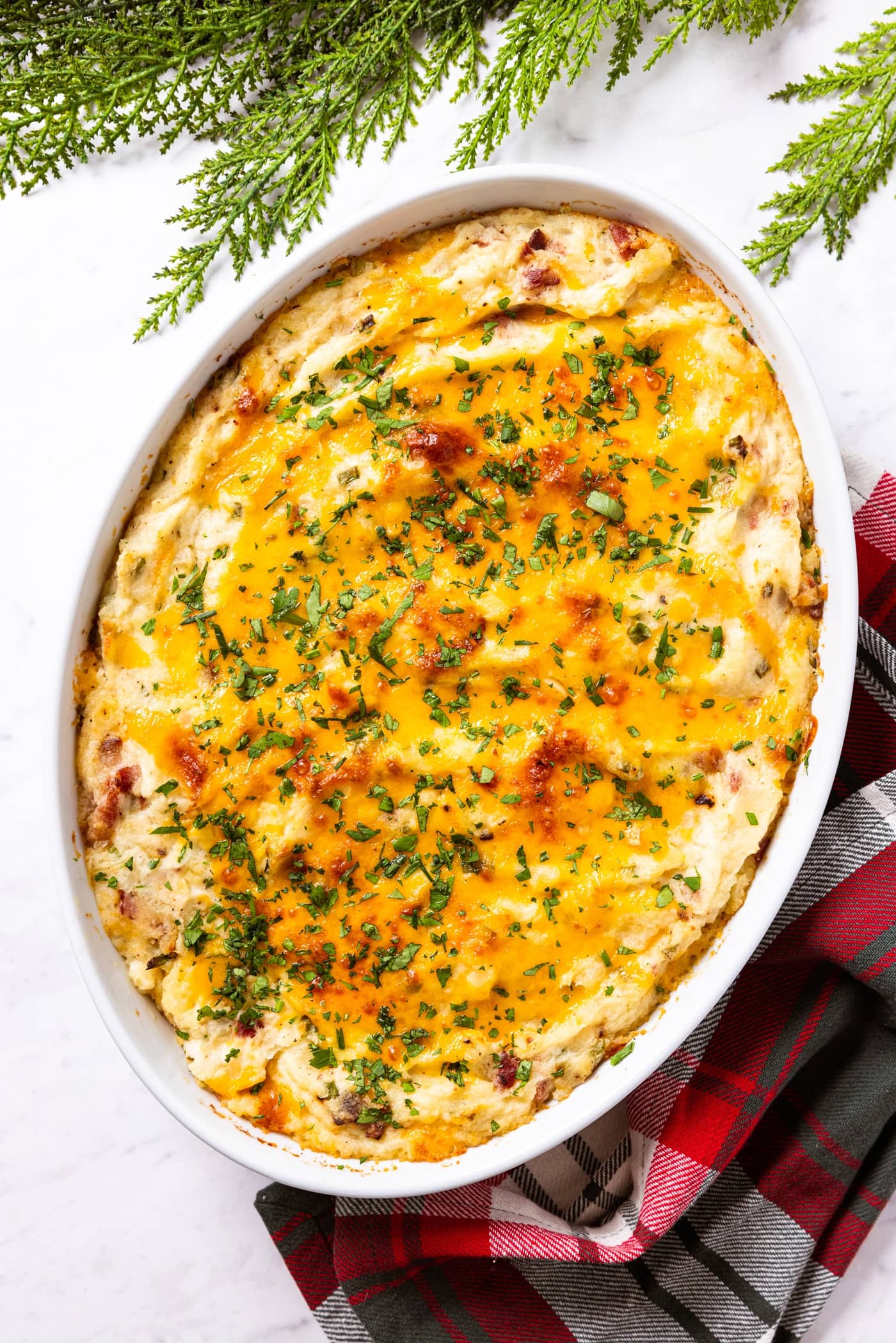 White marble surface with white oval baking dish filled with potato casserole with browned cheese on top with red and green napkin underneath as well as green garland along the top.
