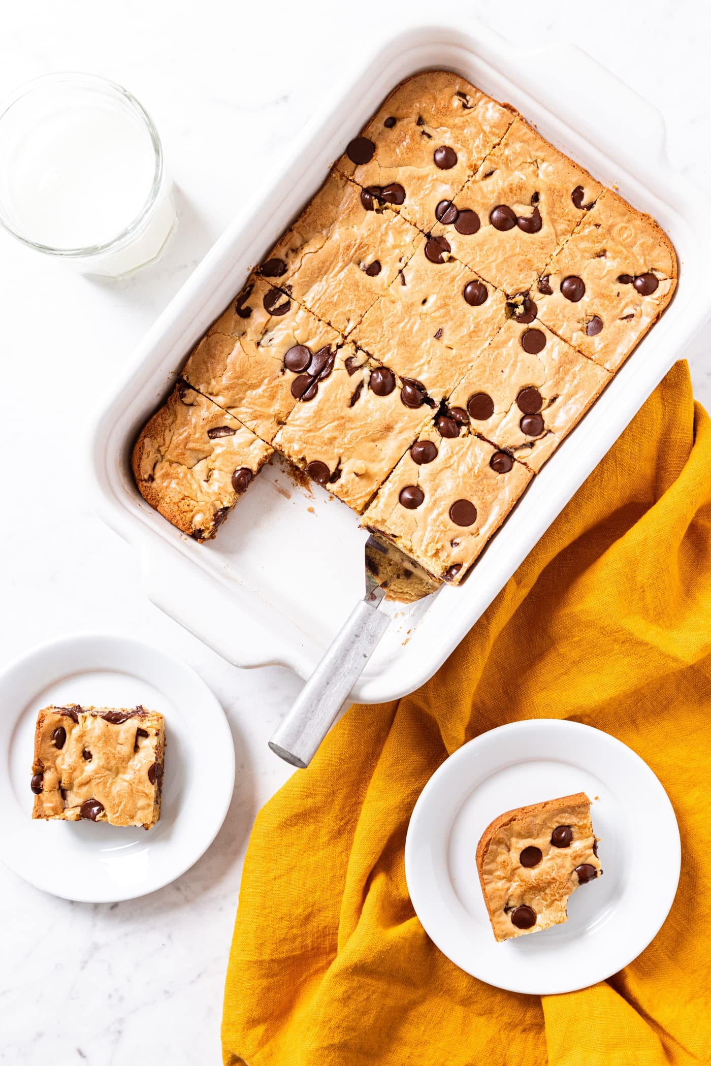 White marble surface with white baking dish filled with baked blondies with two pieces of blondies on small white palates with glass of milk.
