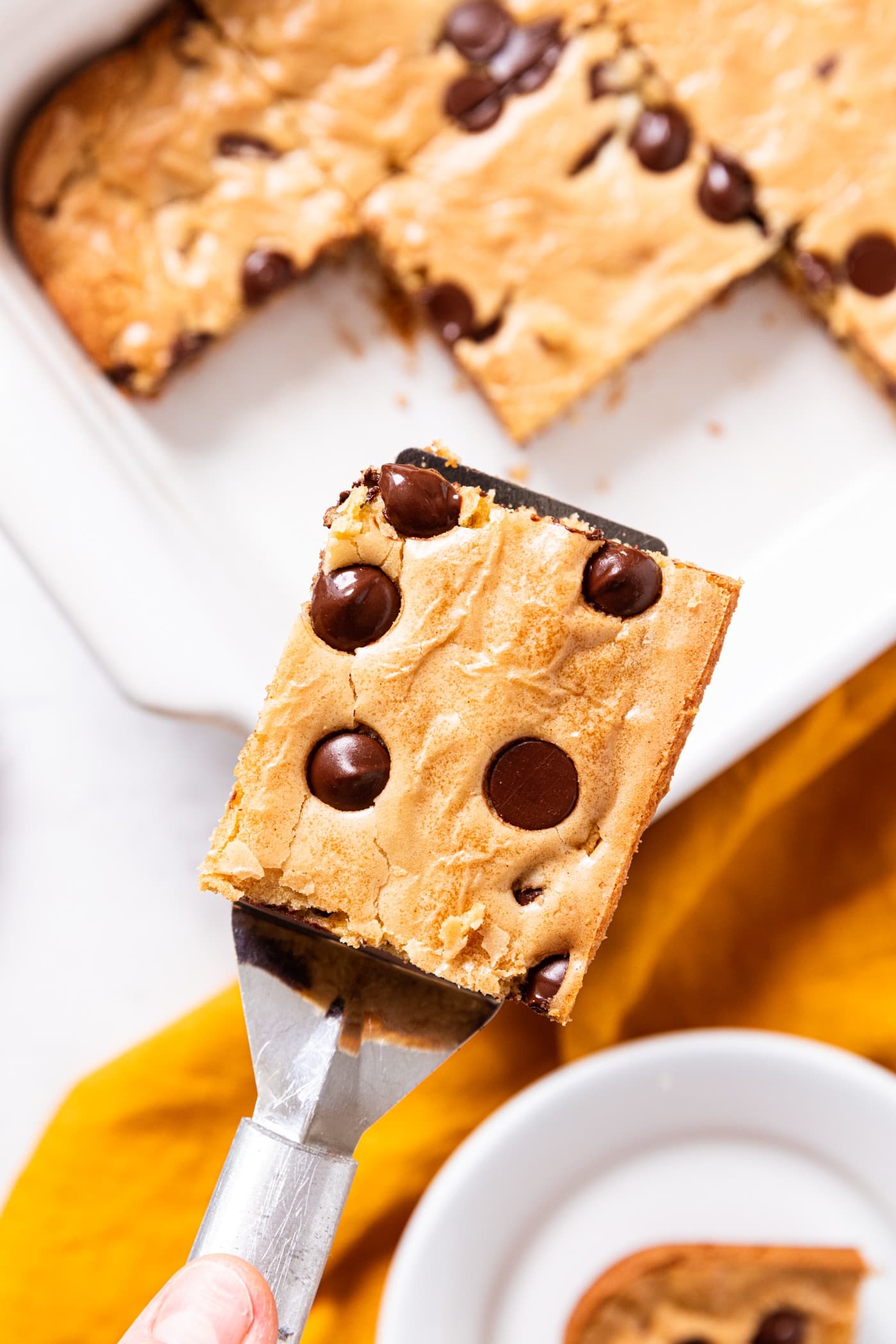 Metal spatula holding serving of blondie with chocolate chips dotting the top with rest of blondies below in background with yellow napkin.