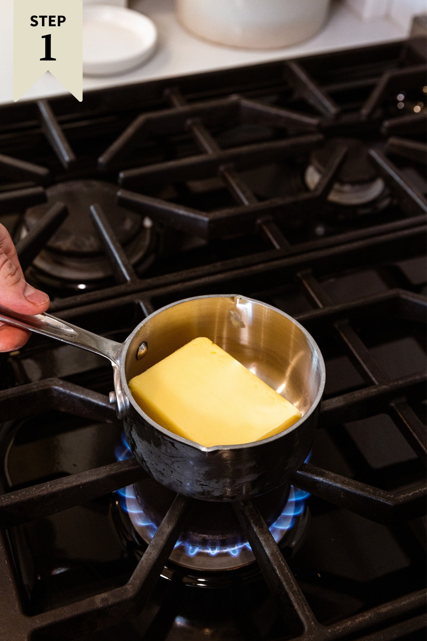 Hand placing small saucepan filled with solid butter before it begins to melt on stovetop.