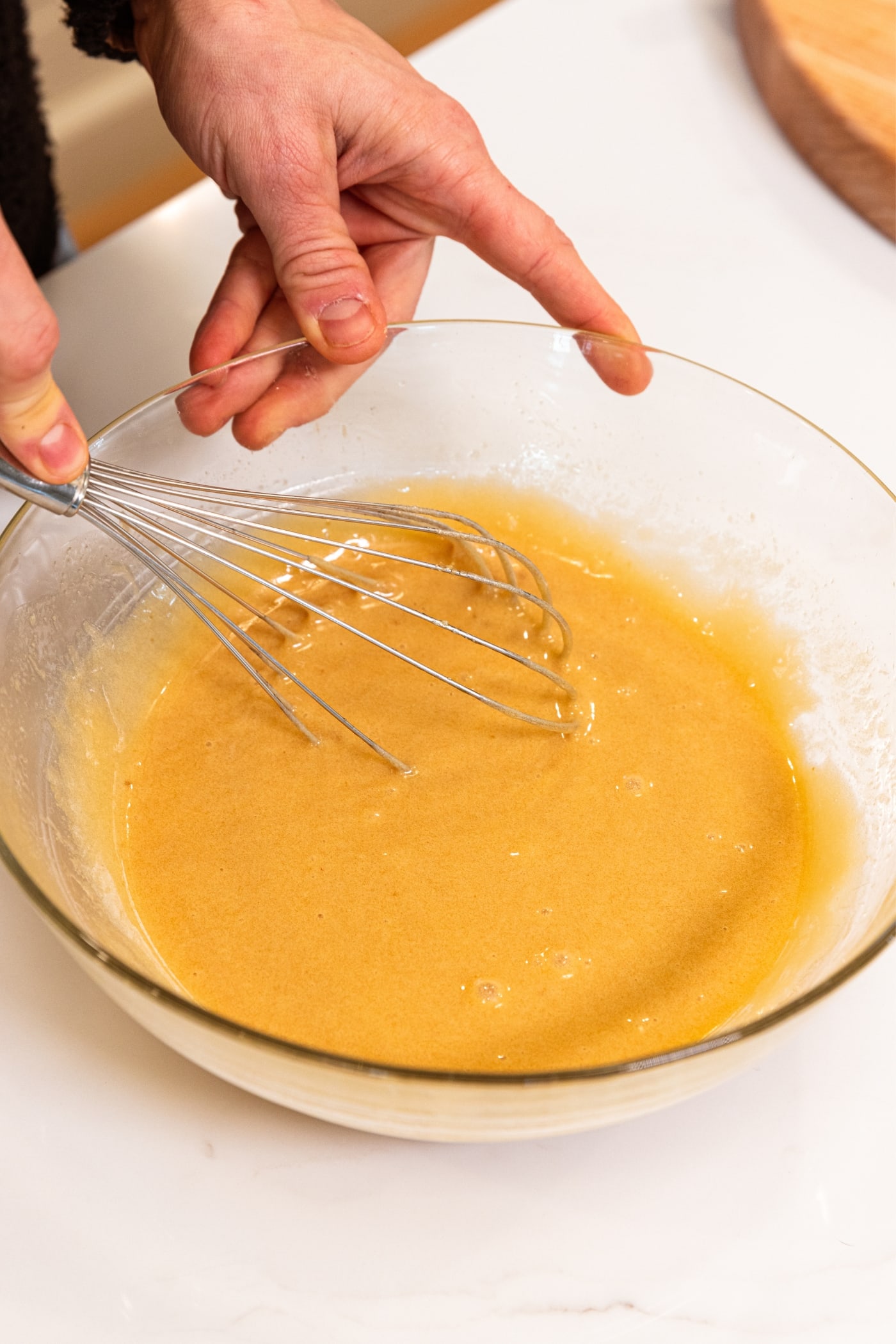 Hand holding whisk whisking together liquid ingredients for classic blondies recipe in large glass bowl on white countertop.