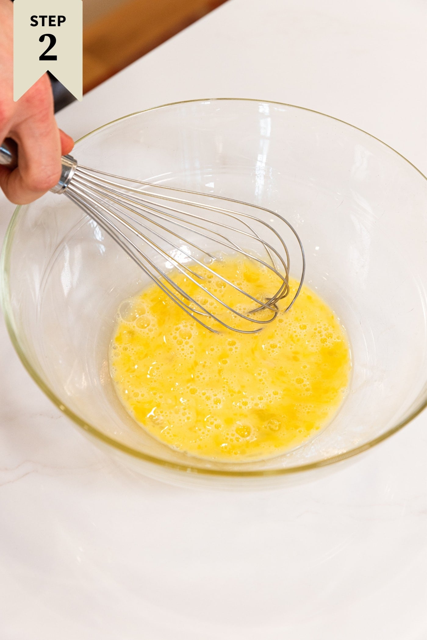 Hand whisking together raw eggs in large glass bowl on white countertop before putting in all of the rest of the ingredients in recipe.