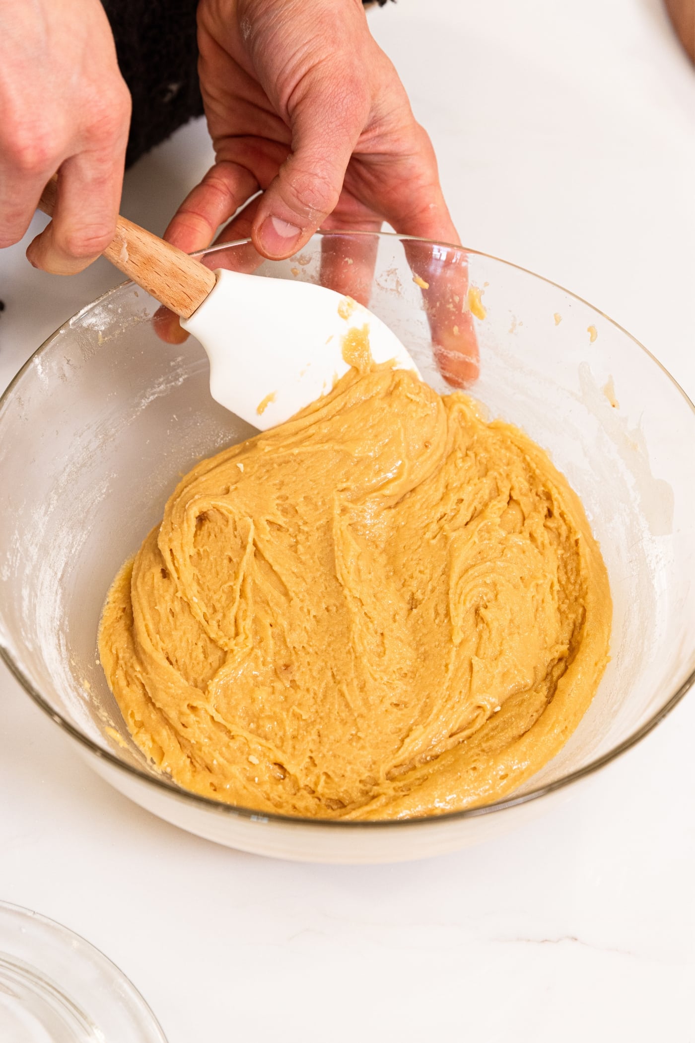 Hand holding spatula finishing mixing together thick brown-colored batter for blondies in glass bowl on white countertop.