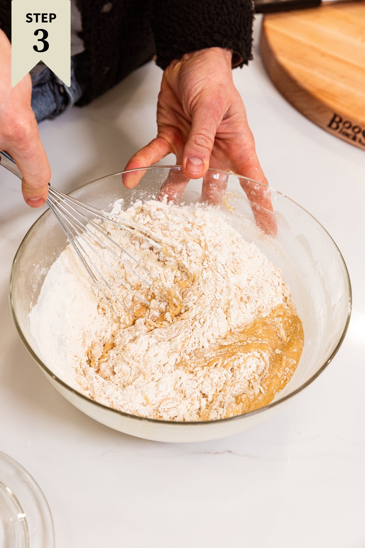 Hand whisking together dry flour into wet ingredients for classic blondies recipe in large glass bowl on white countertop with wood cutting board in background.