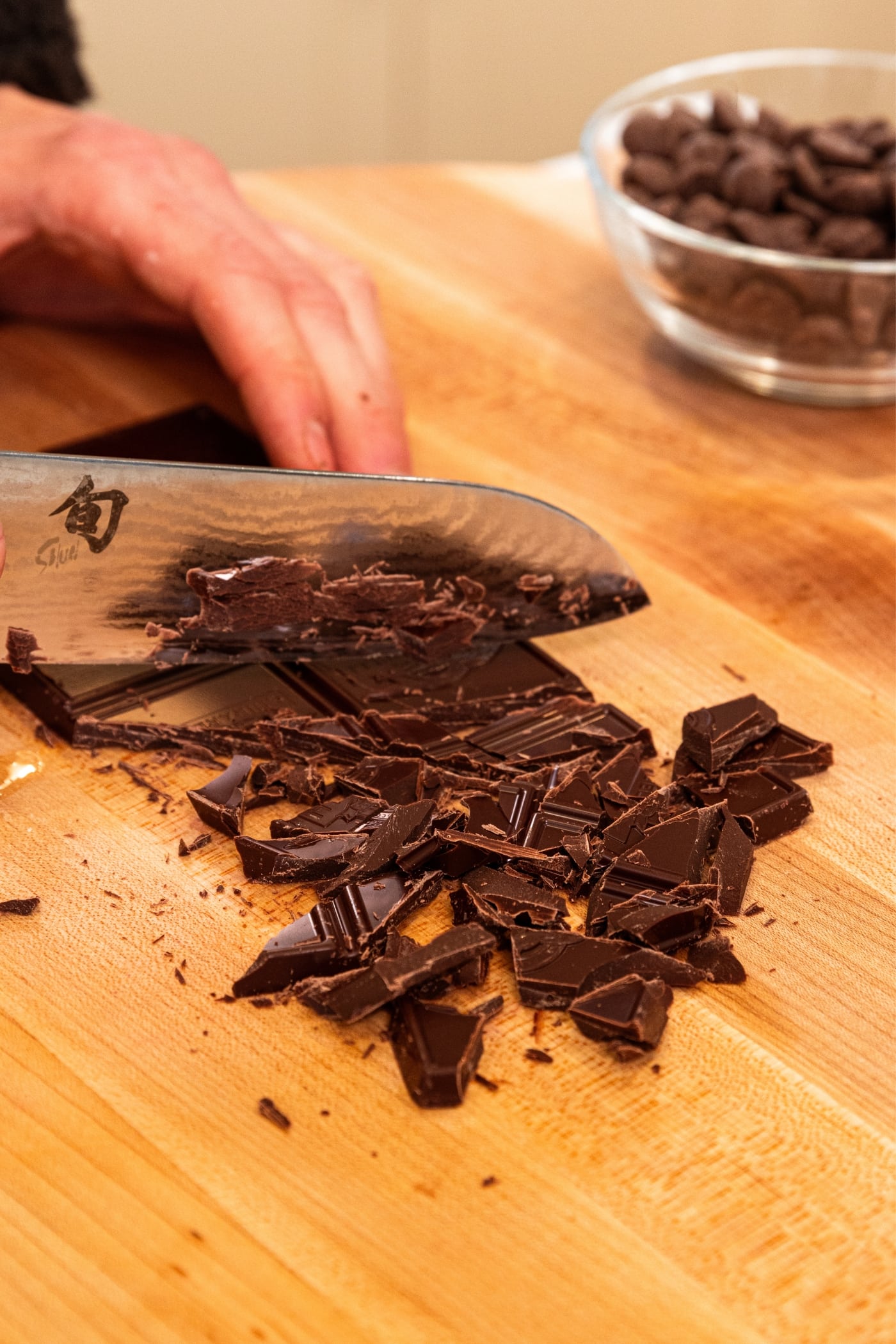 Hand holding knife slicing up shards of chocolate from chocolate bar on wood cutting board.