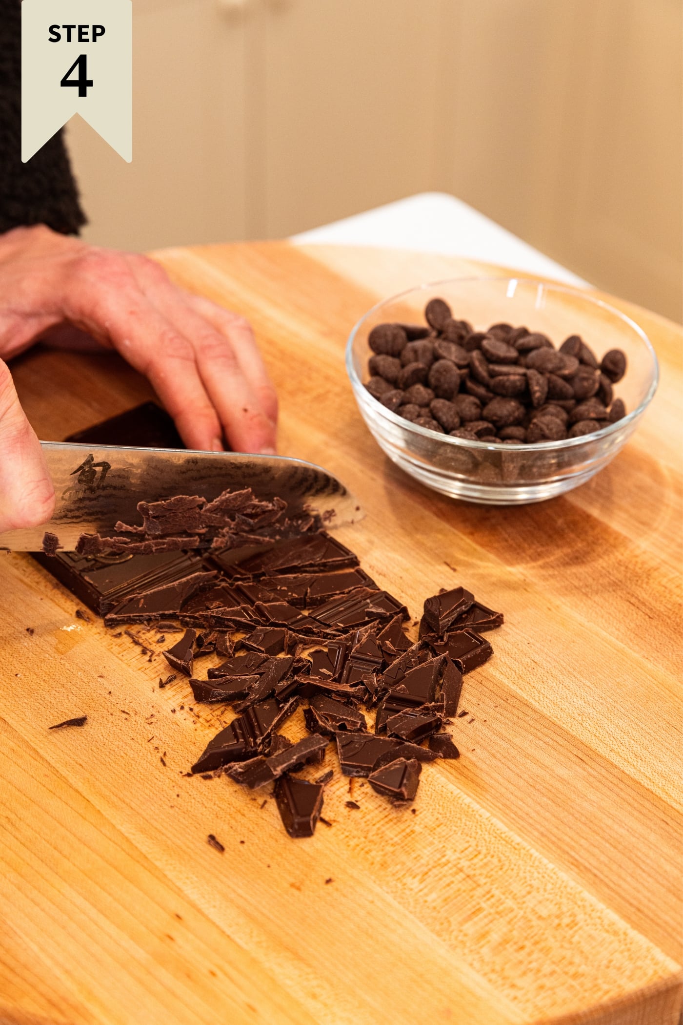 Hand using knife to chop up shards of chocolate from chocolate bar on wood cutting board with small glass bowl filled with chocolate chips.