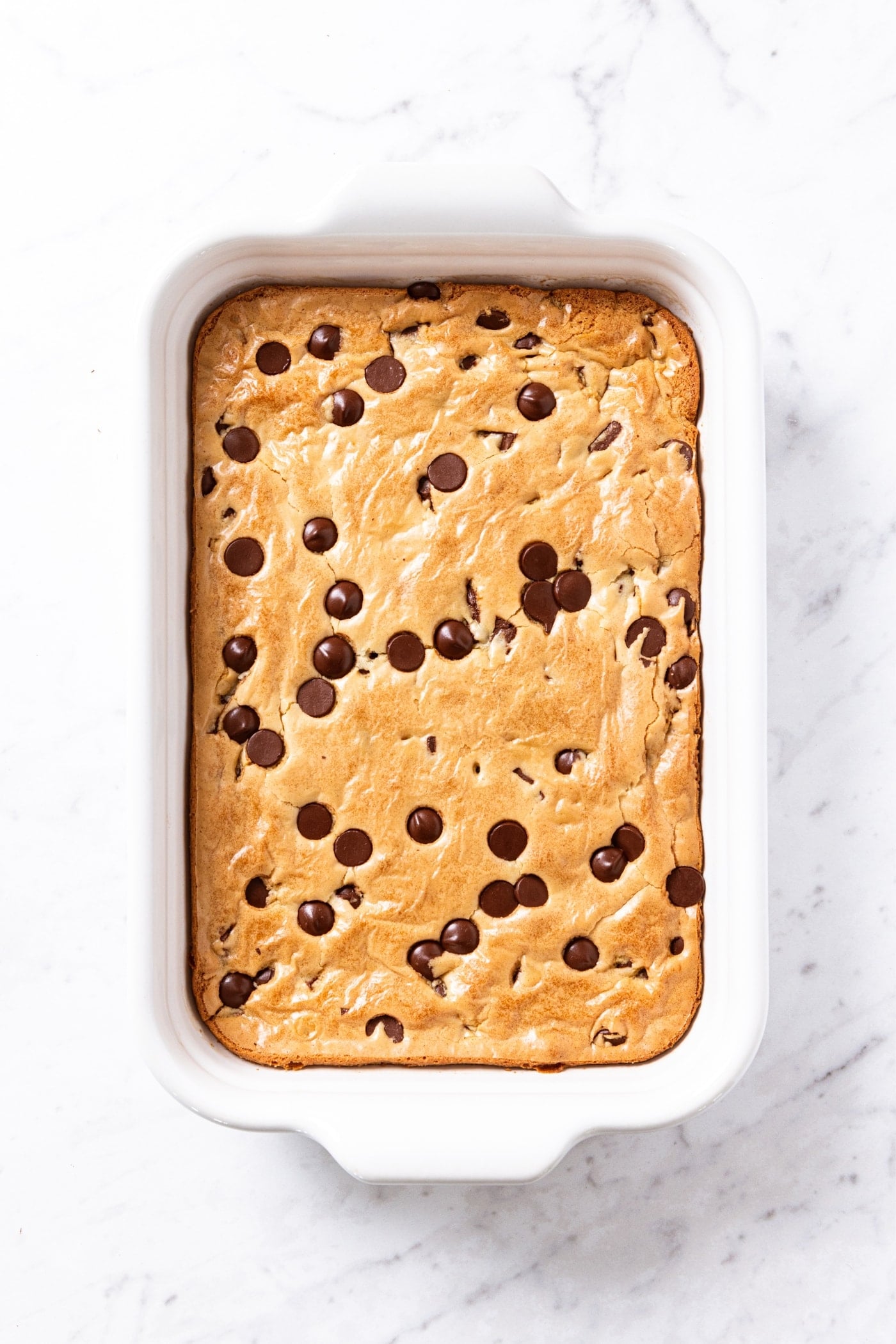 White baking dish sitting on white marble surface with freshly baked blondies in pan with chocolate chips all over the top but with blondie texture showing through.