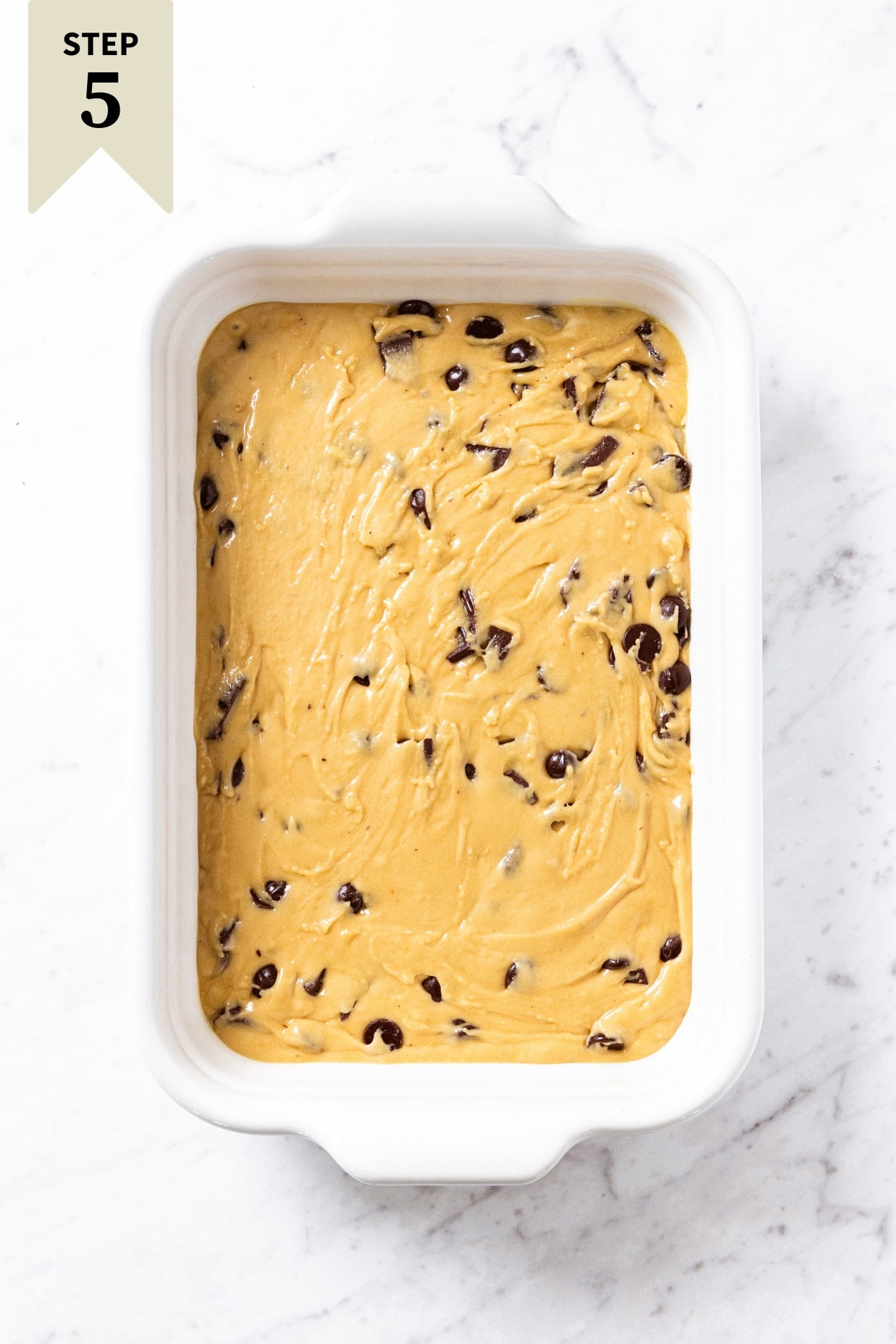 White marble surface with 9x13 baking dish filled with unbaked batter for blondies with chocolate chips and chocolate pieces dotting the top right before going into the oven.