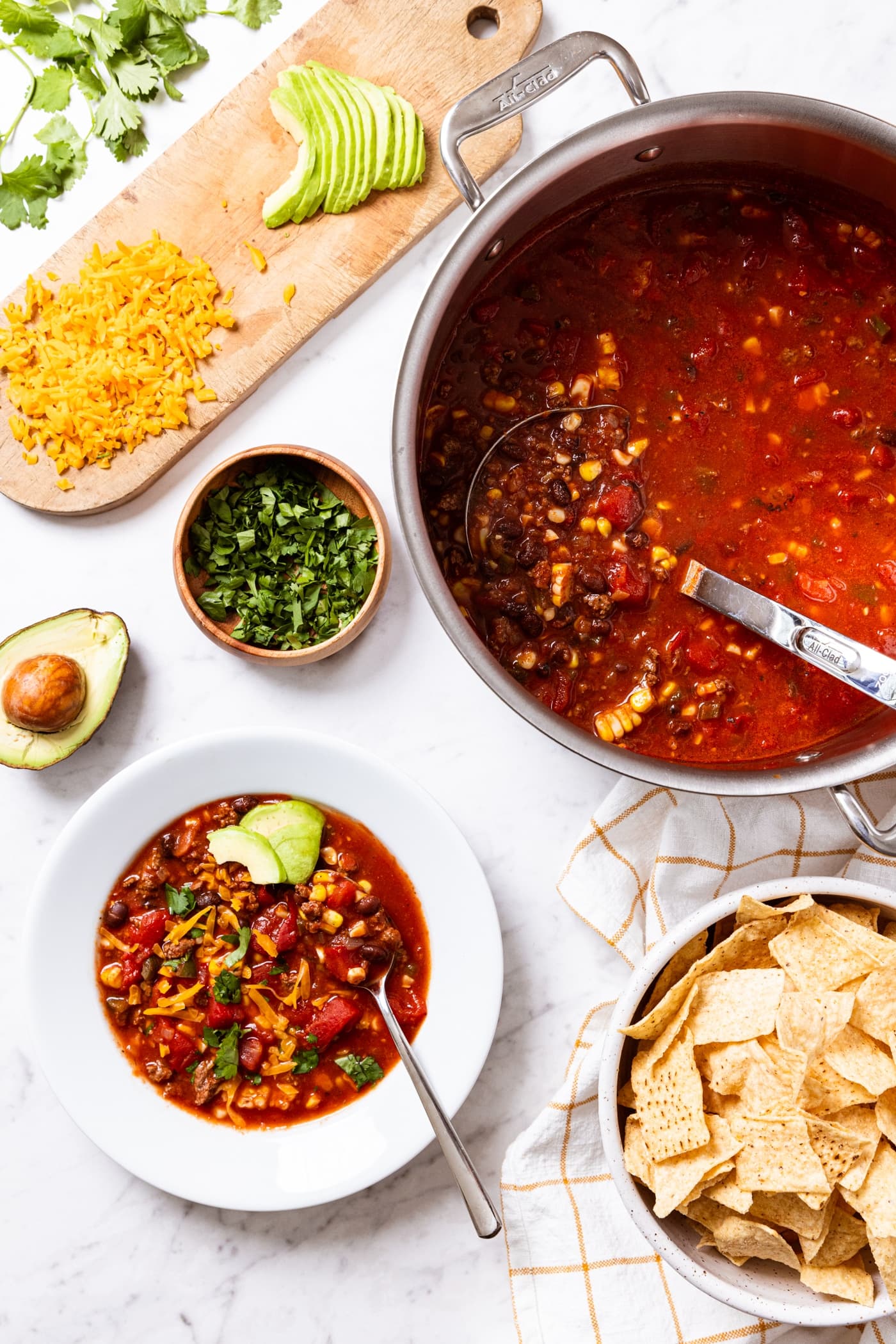 White marble surface with all components of an easy taco soup including hot red colored taco soup and white bowl filled with serving of soup fresh from the stockpot with toppings all spread around to put on top of the soup.