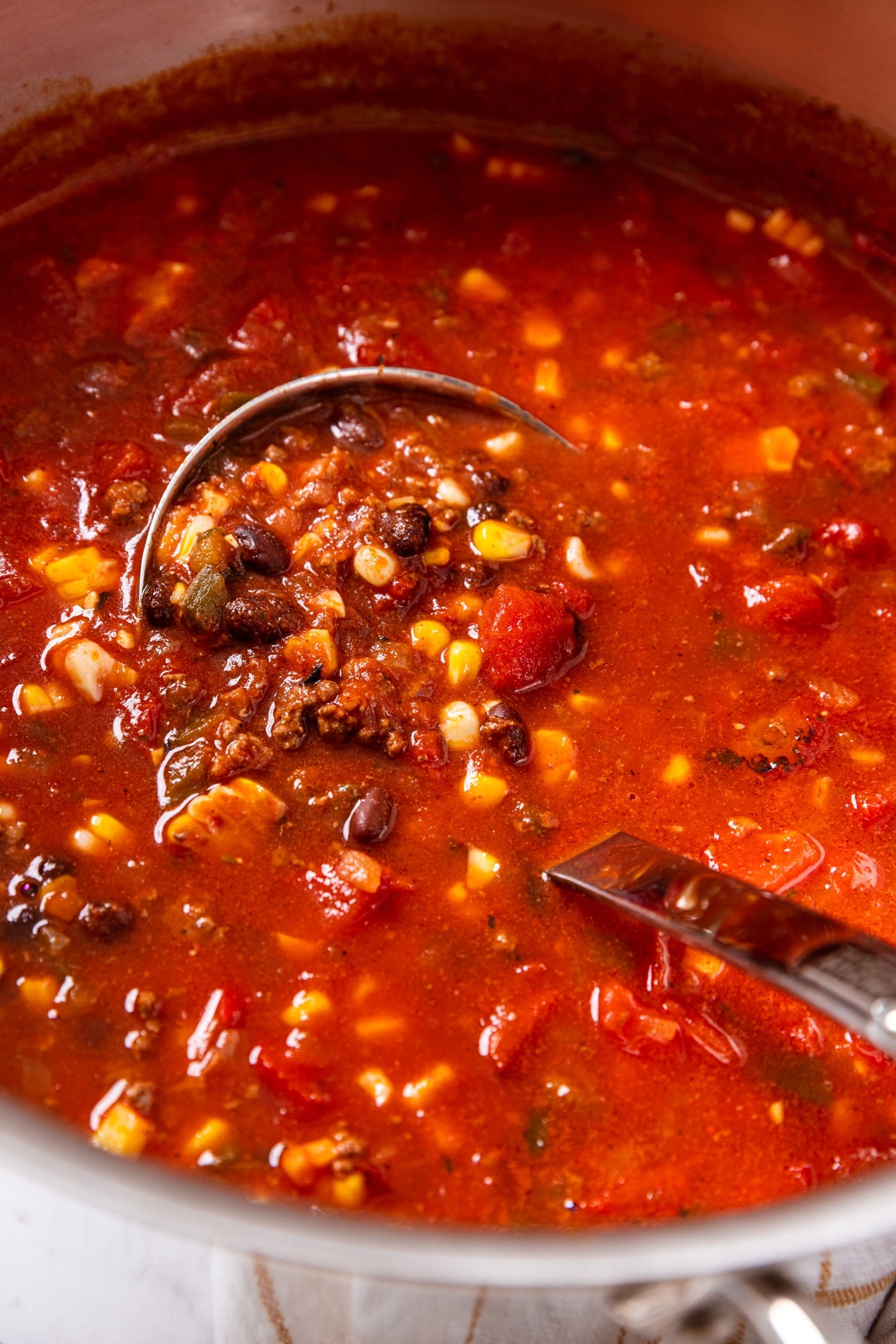 Metal stockpot filled with red-colored taco soup with pieces of corn, black beans, and browned beef showing as portion is held in metal ladle.