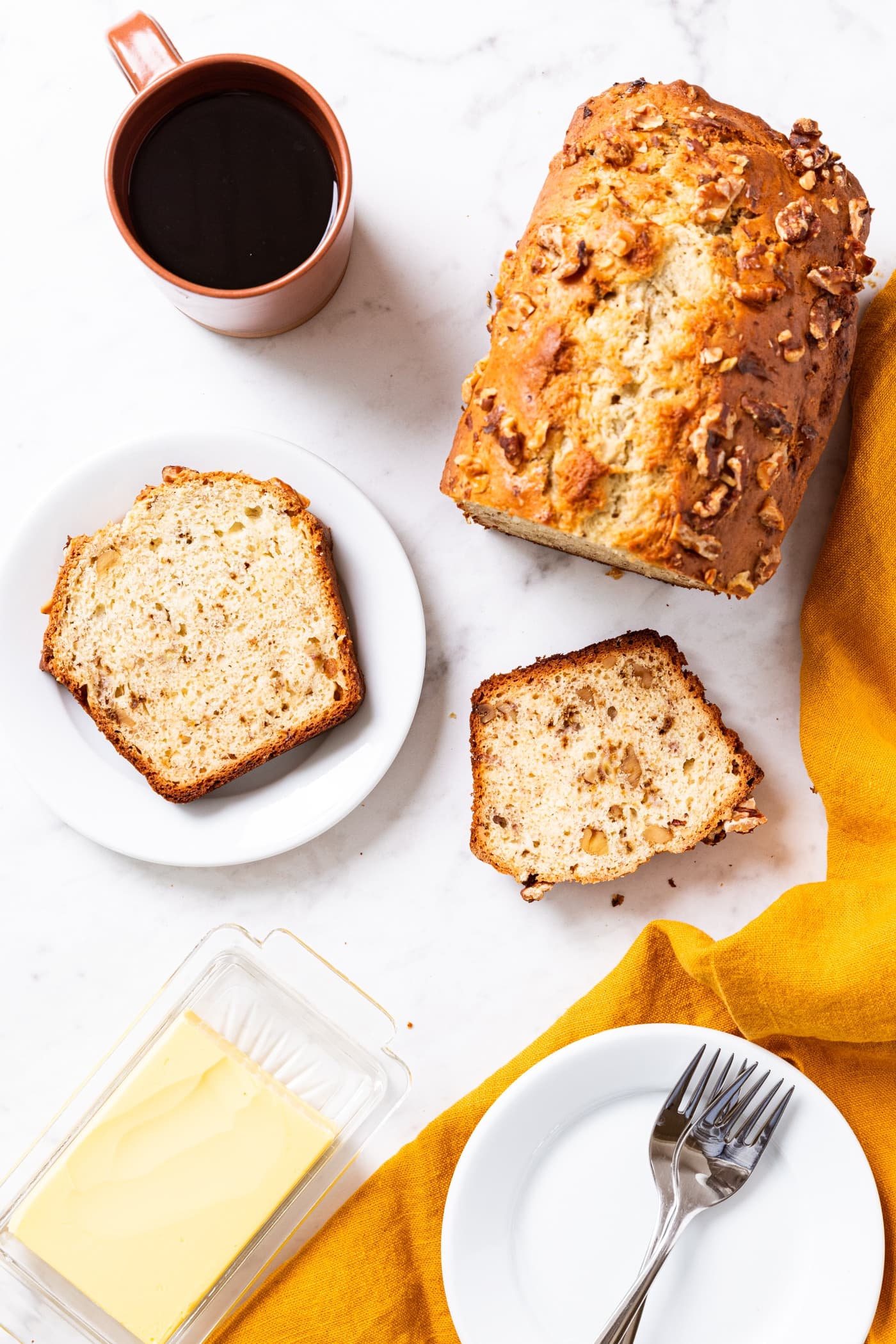 White marble surface with banana bread slices sitting on surface along with white plate with orange coffee mug filled with black coffee.