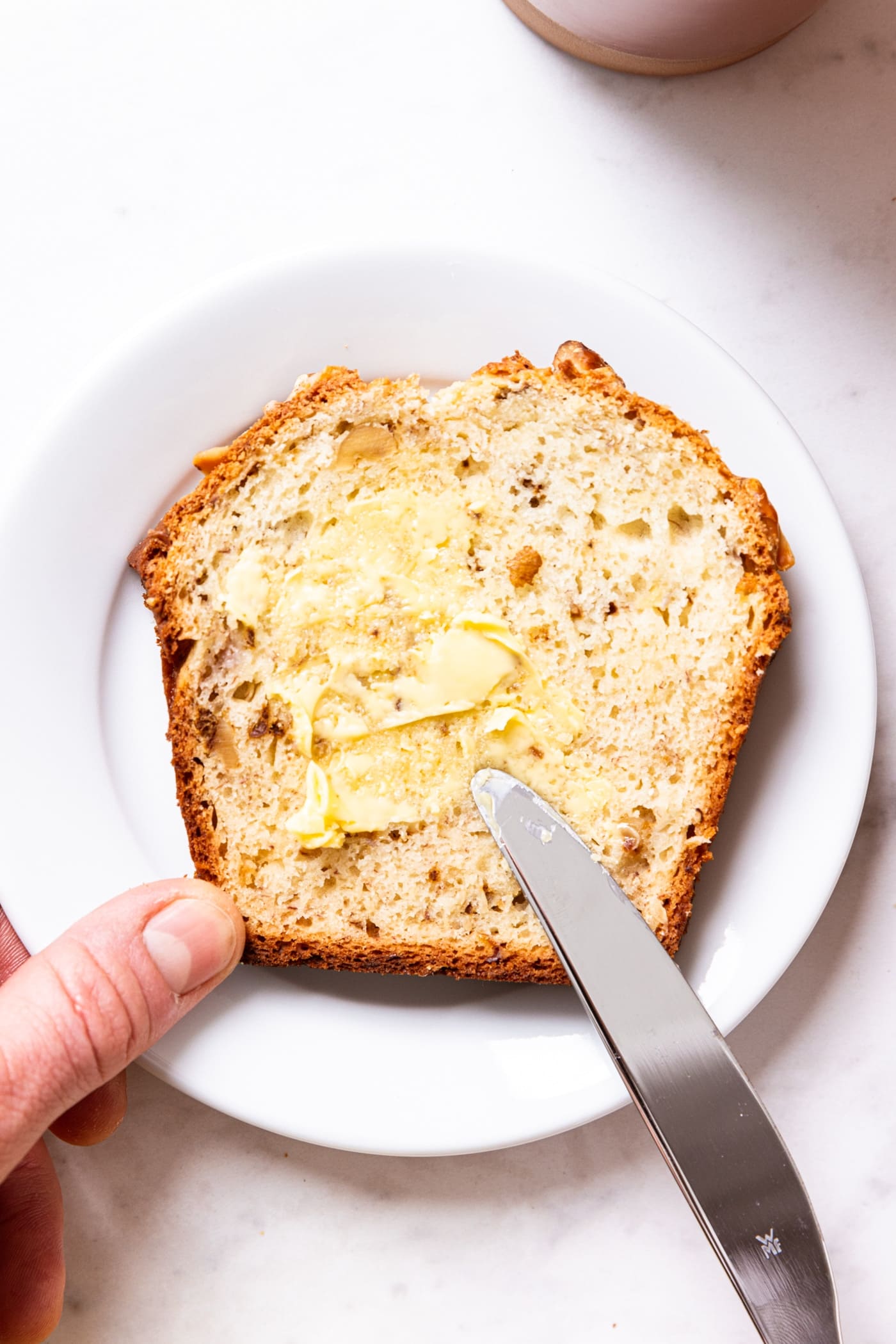 White plate with slice of classic banana bread with hand using knife to spread butter on surface of bread showing walnuts in interior of bread.