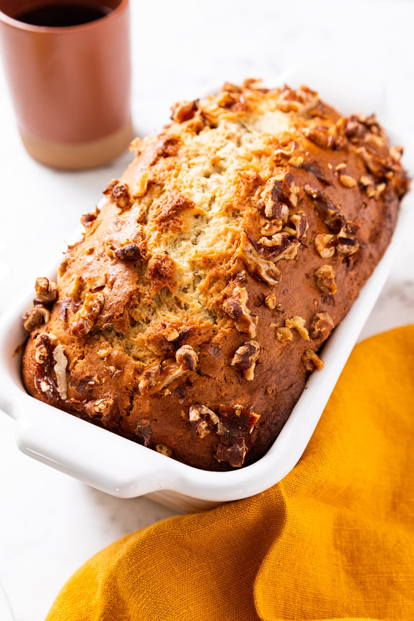 Baked loaf of banana bread with browned top with walnuts sitting on white marble surface with orange coffee mug in background and yellow napkin to the side.