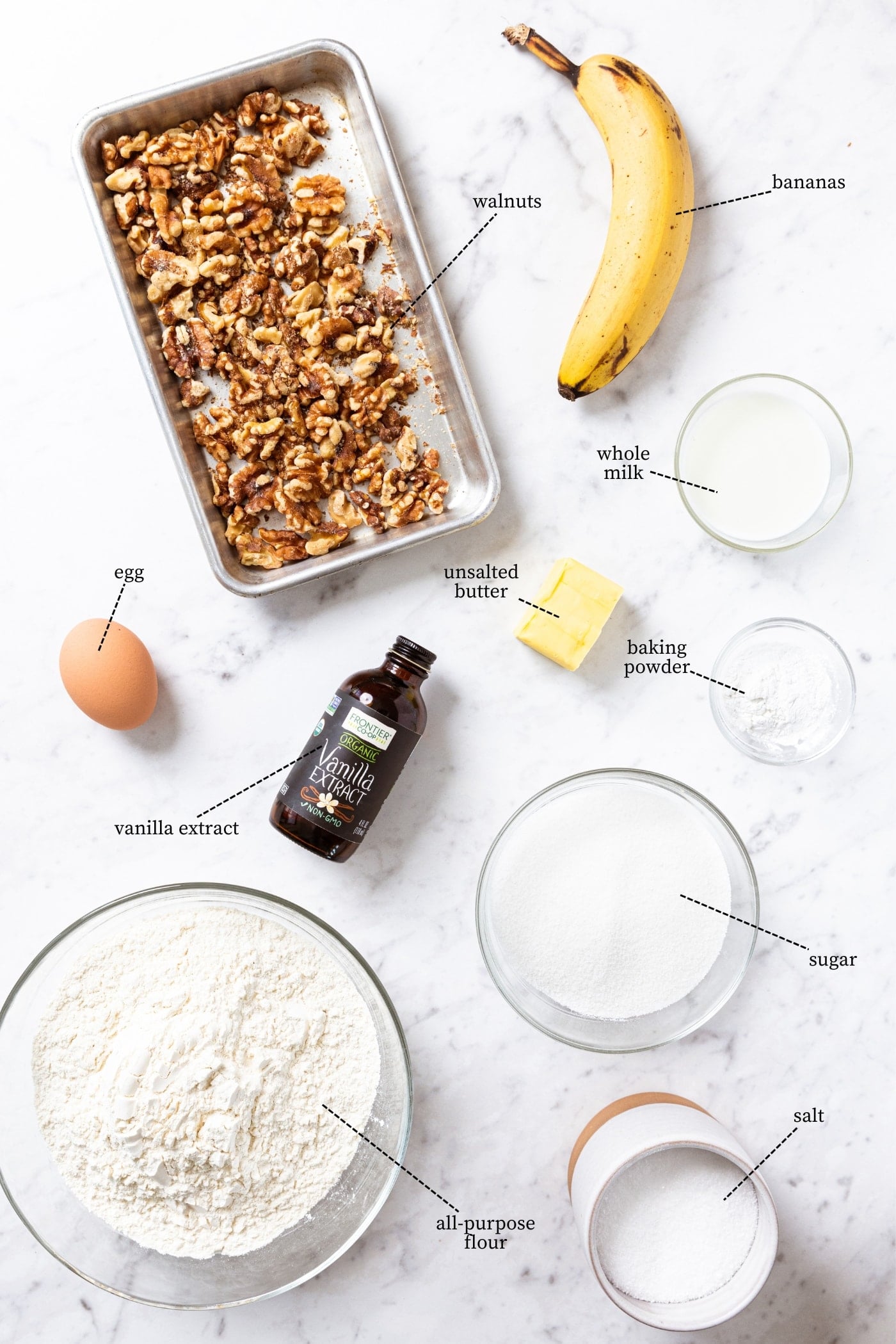 White marble surface filled with all ingredients needed to make classic banana bread including flour, sugar, walnuts, banana, spices, and more.