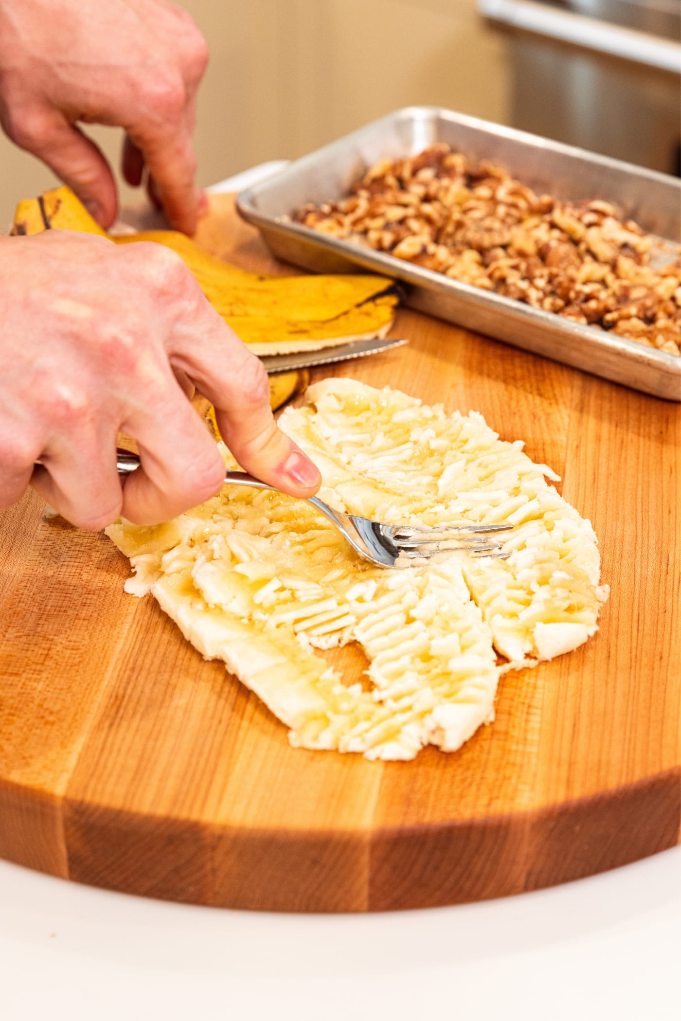 Hand using fork to mash bananas into a smashed texture on wood cutting board with walnuts on metal tray in background.