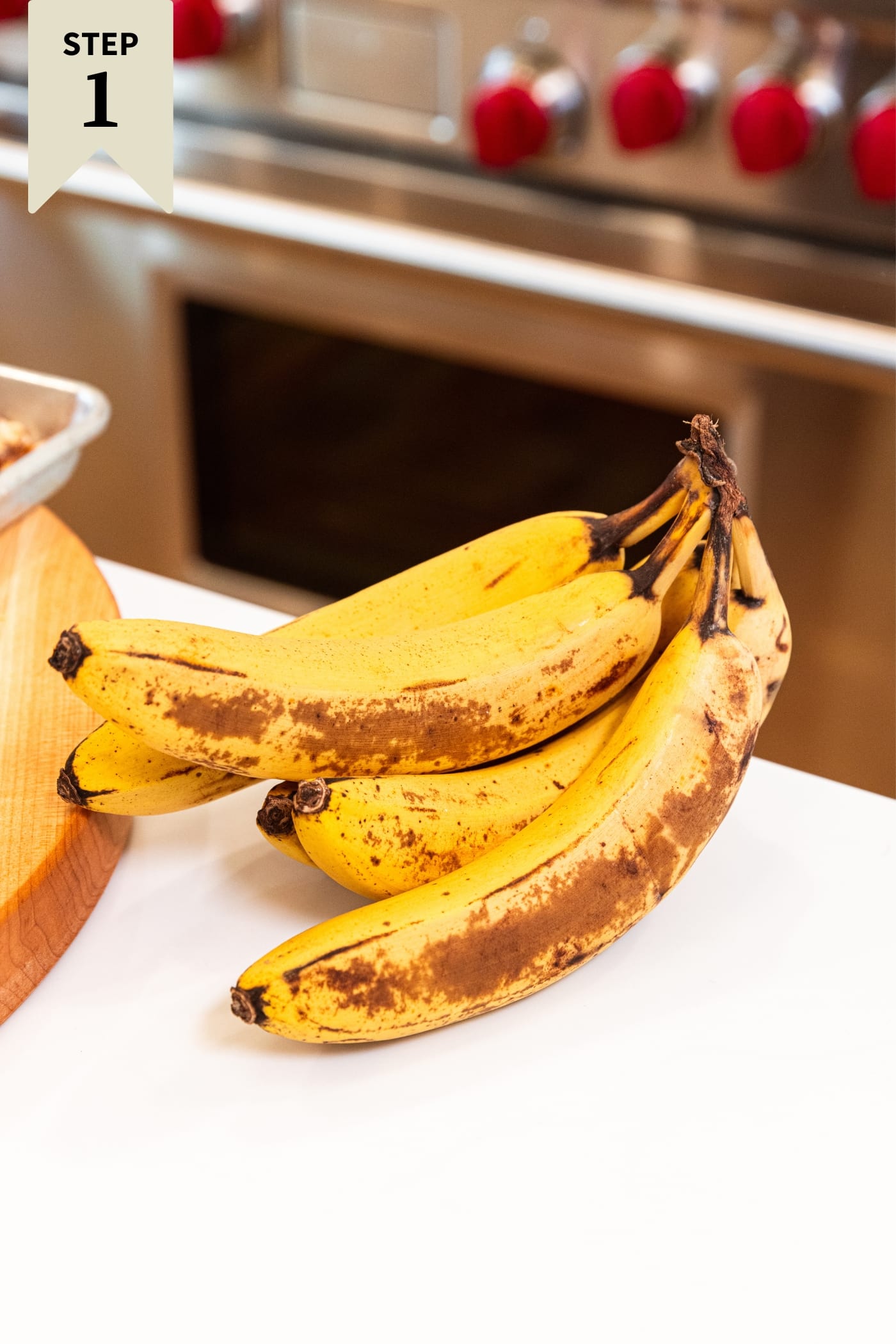 White surface with brown bananas still in a bunch sitting on countertop with stainless steel oven in background.