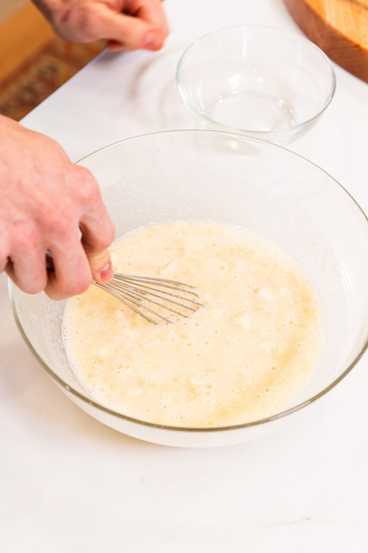 Hand holding whisk mixing together mashed banana with milk and sugar in glass bowl on white surface.