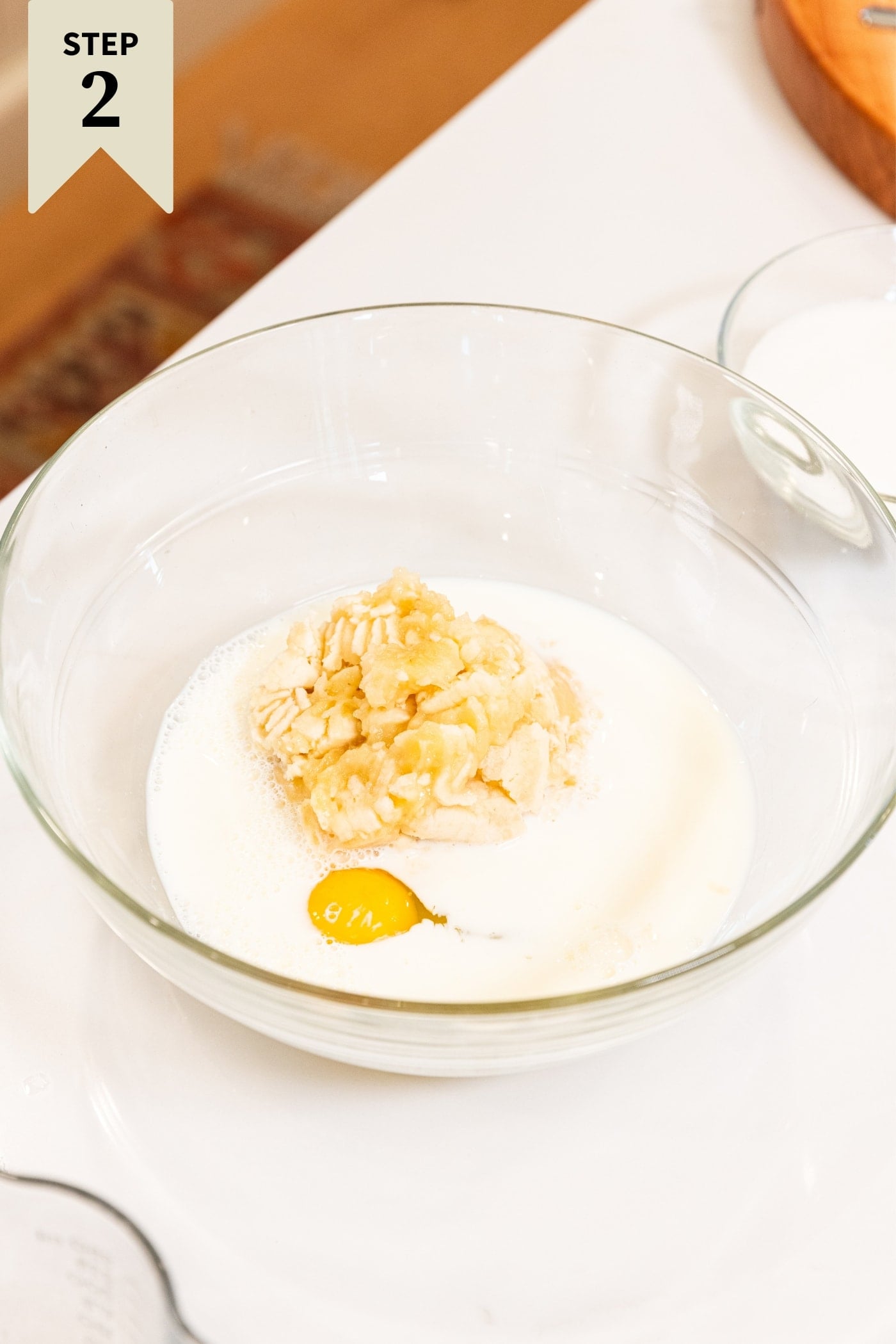 Glass bowl filled with milk, mashed bananas, and an egg sitting on white surface before being mixed together.
