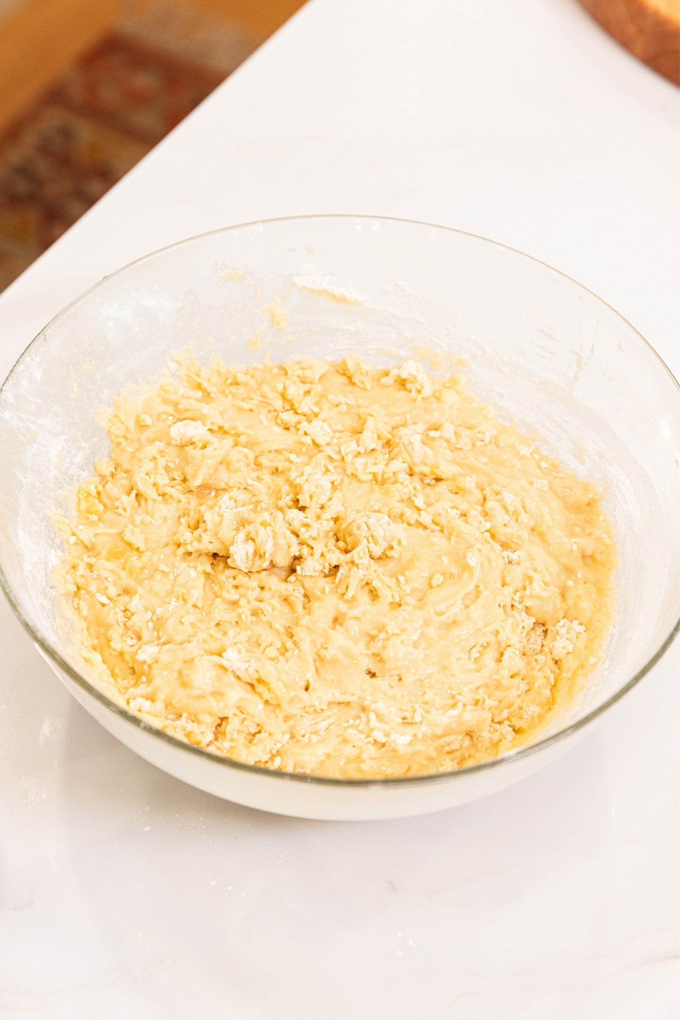 Glass bowl filled with roughly mixed batter for making banana bread all on white surface.