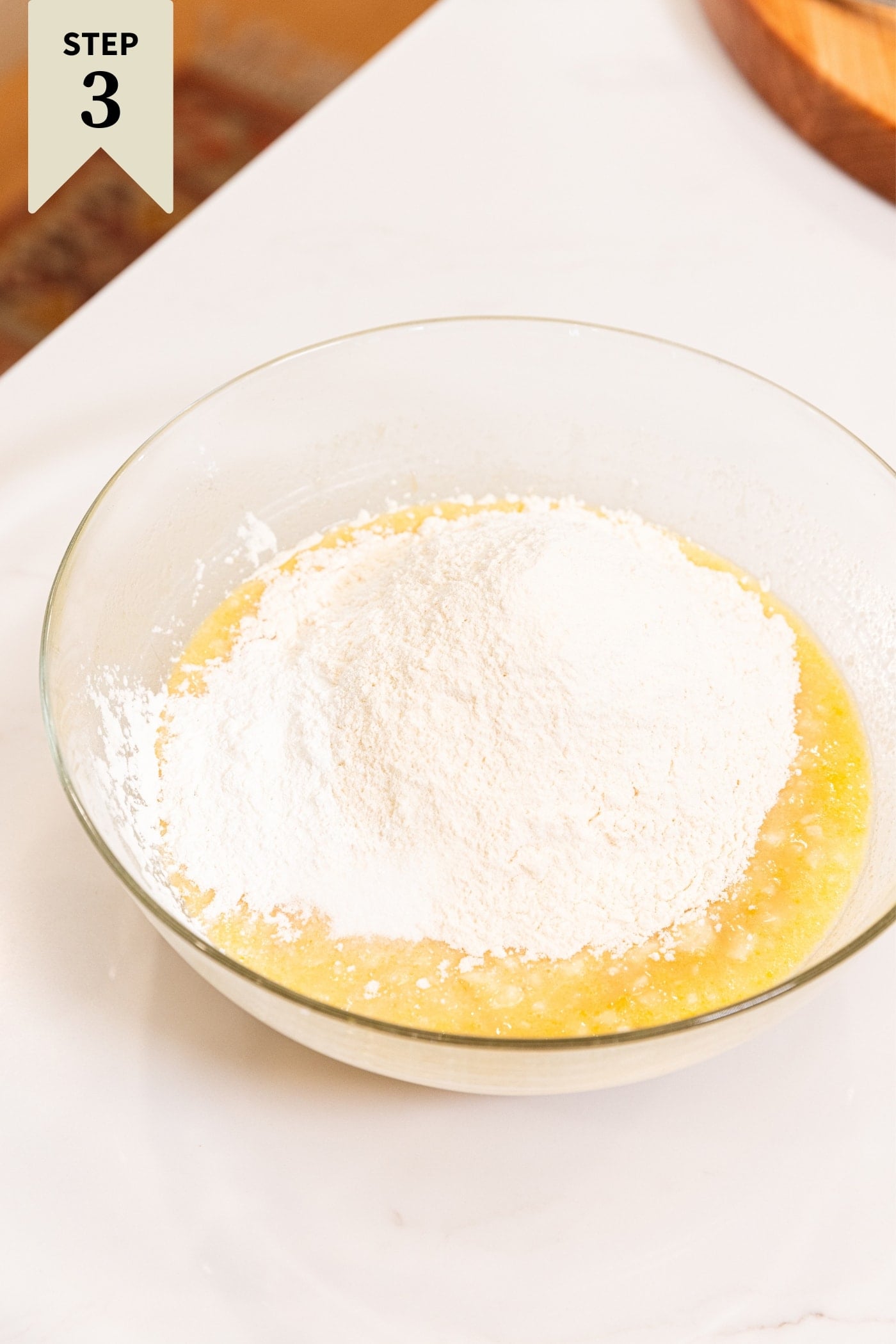 White surface with glass bowl with flour poured on top of yellow colored batter before being mixed together.