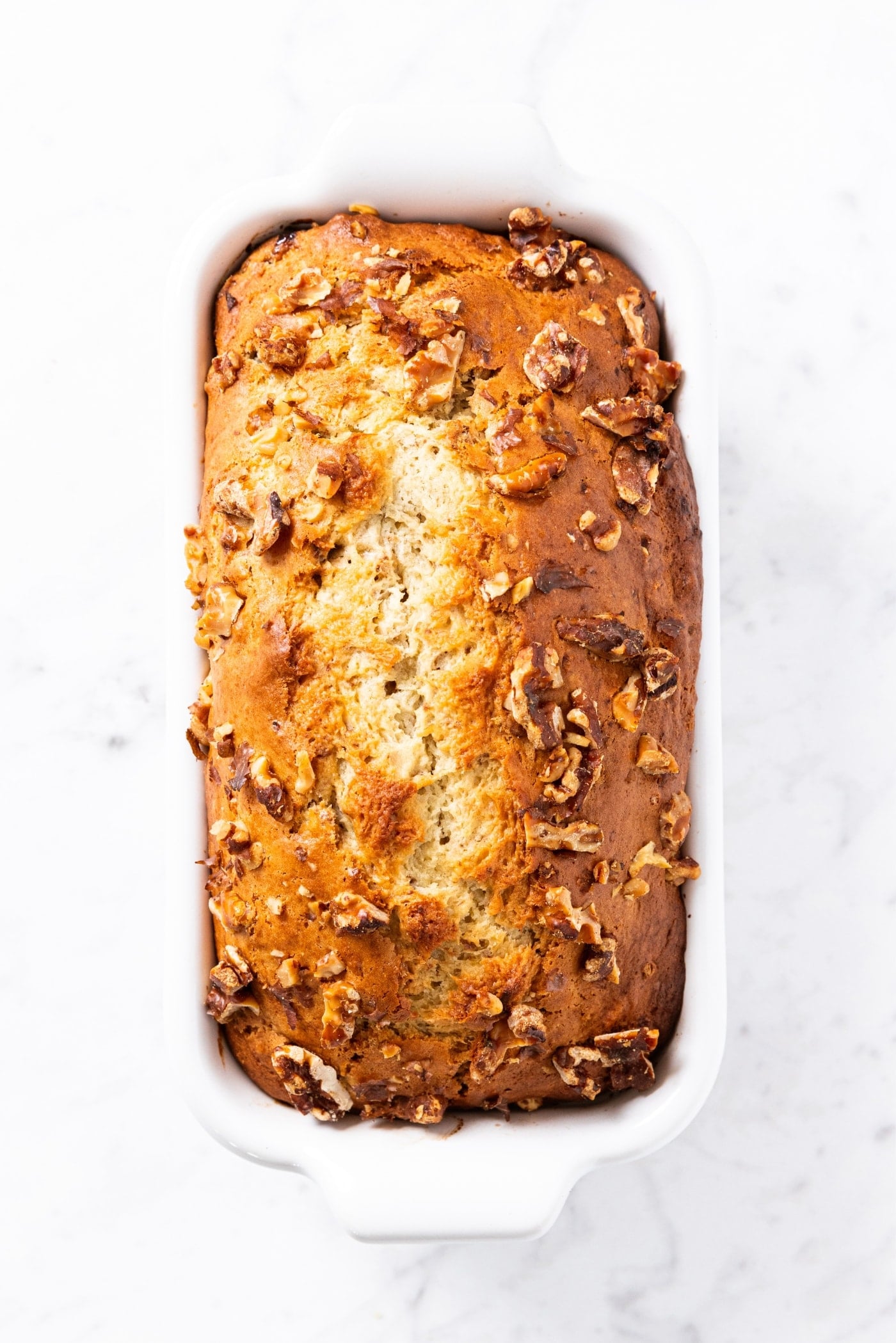 Baked loaf of banana bread sitting on marble surface after coming out of oven with brown crispy top with nuts all over the top.