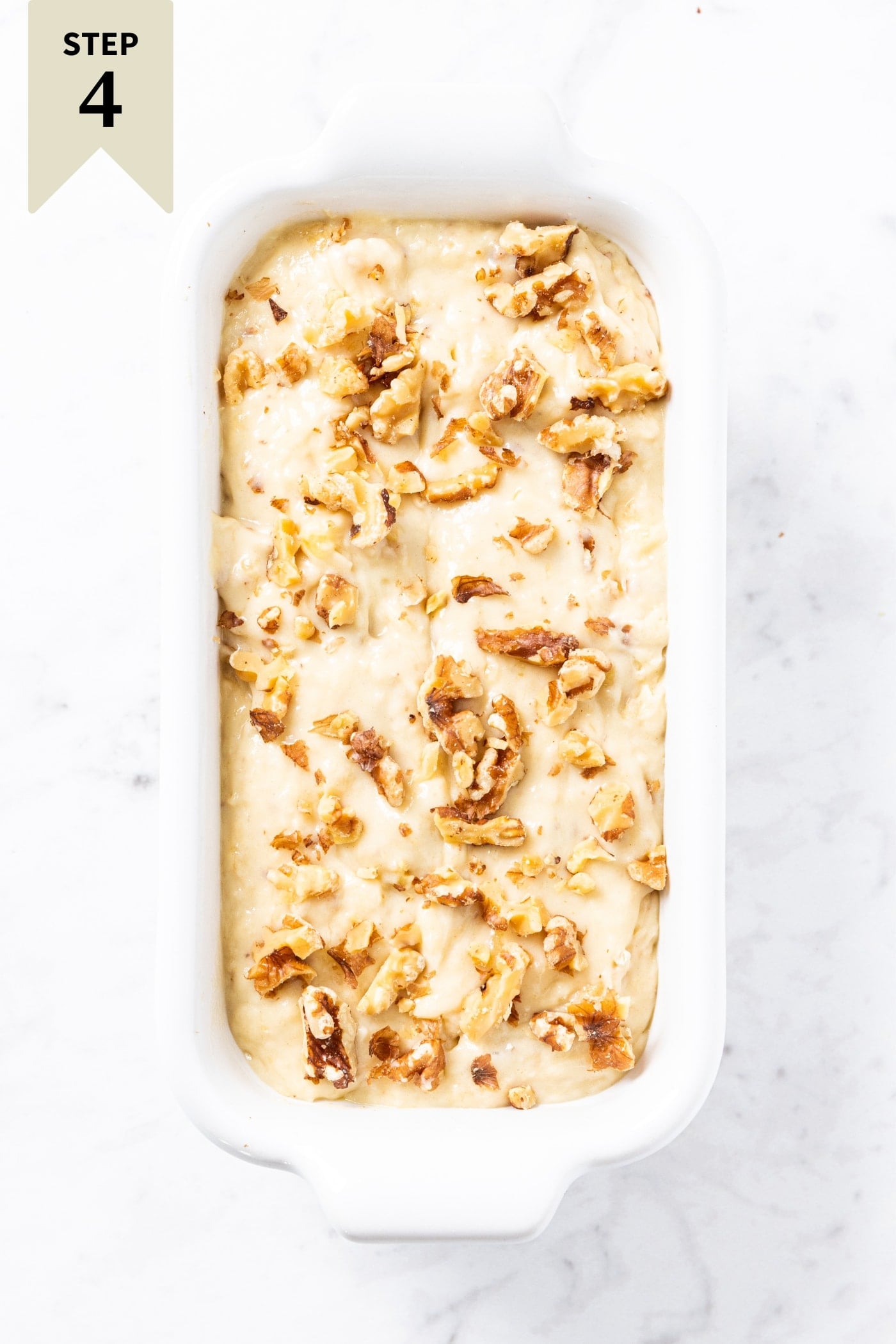 White baking dish filled with unbaked banana bread with walnuts sprinkled on top sitting on white marble surface.
