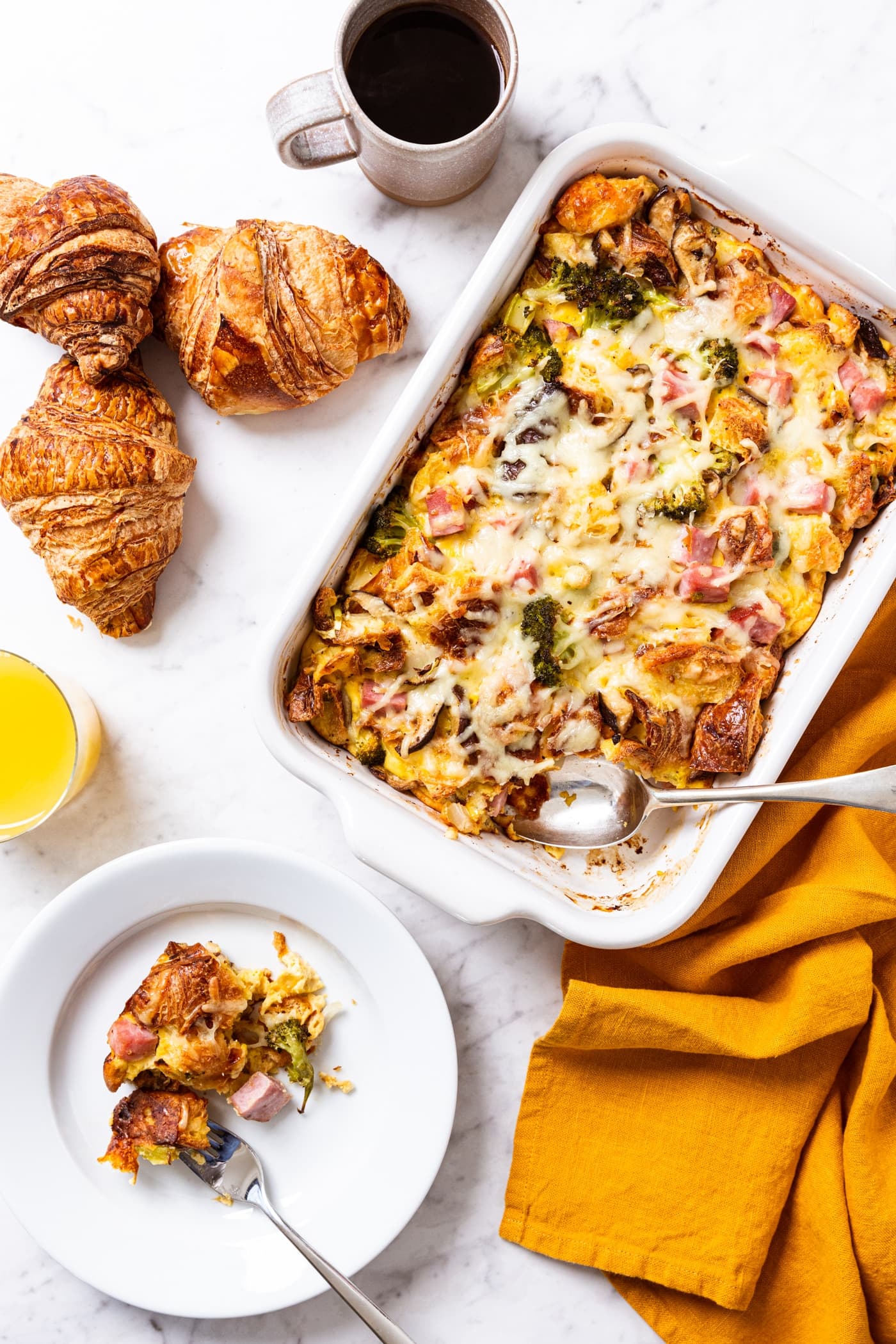 White marble surface with white baking dish filled with baked egg casserole inside with croissants, orange juice, coffee, and plate of serving of casserole.