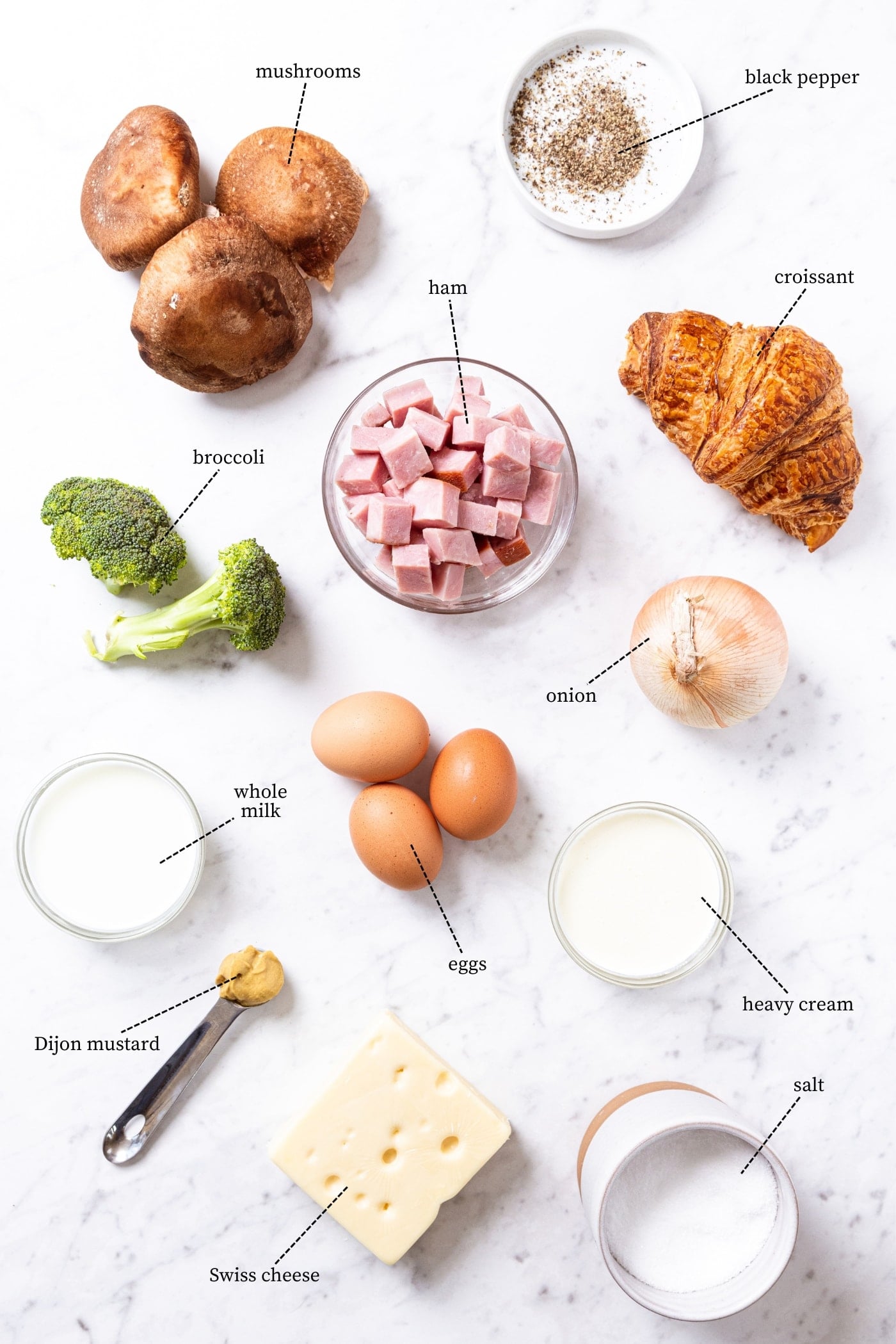 White marble surface filled with all ingredients needed to make croissant egg casserole including eggs, mushrooms, broccoli, cheese, onion, spices, croissant, and more.