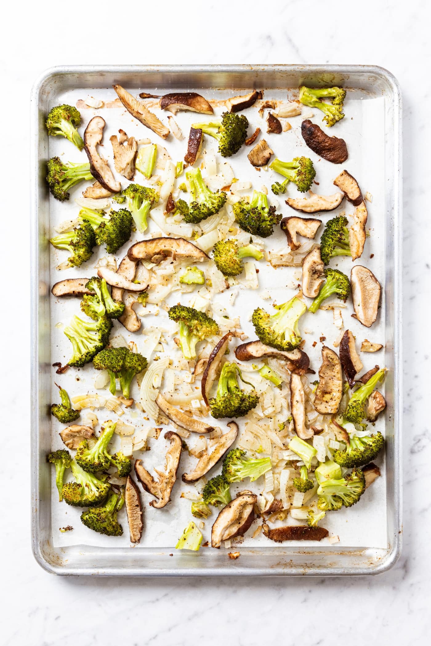 White marble surface with metal baking sheet filled with roasted vegetables after coming out of the oven including broccoli, mushroom, and onion.