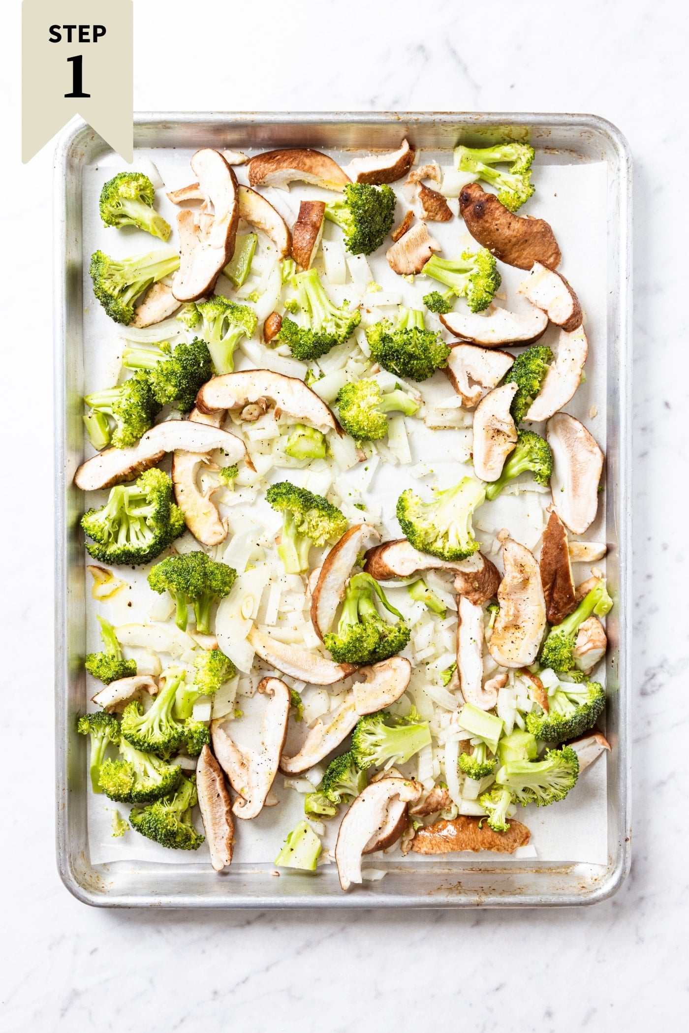 White marble surface with metal baking sheet filled with raw slices of broccoli, mushroom, and onion all doused in olive oil, salt, and black pepper right before going into the oven to roast.