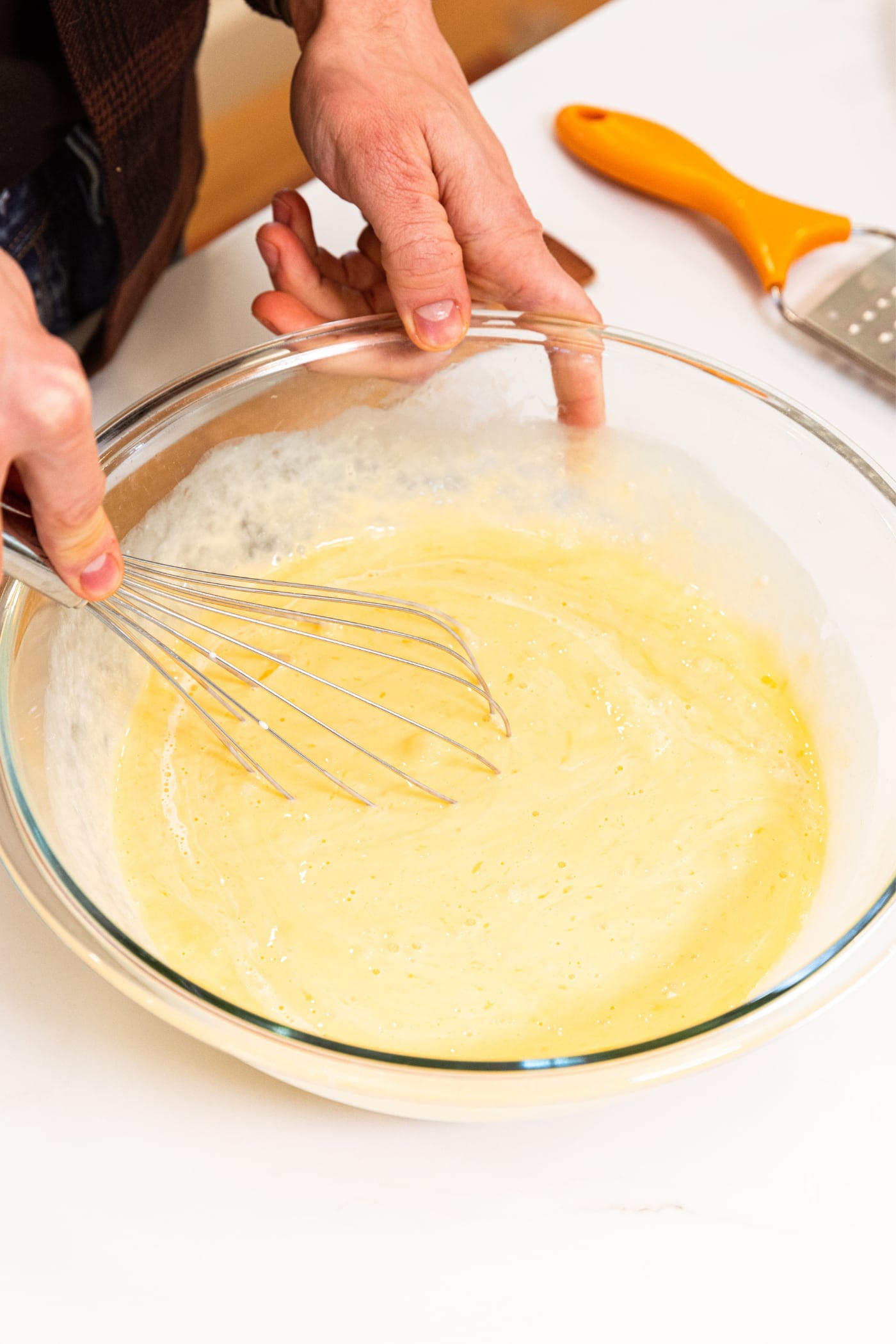 Hand holding whisk mixing together egg casserole mixture in large glass bowl on white countertop.