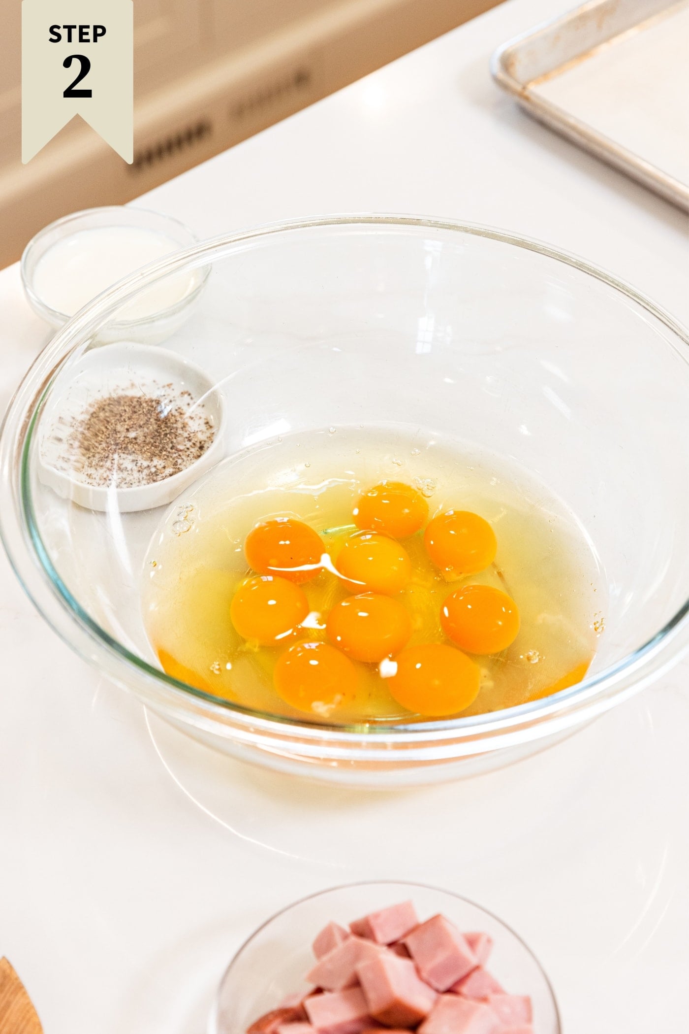 Glass bowl filled with lots of egg yolks and whites sitting on white countertop with small bowl of cubed ham to the front.