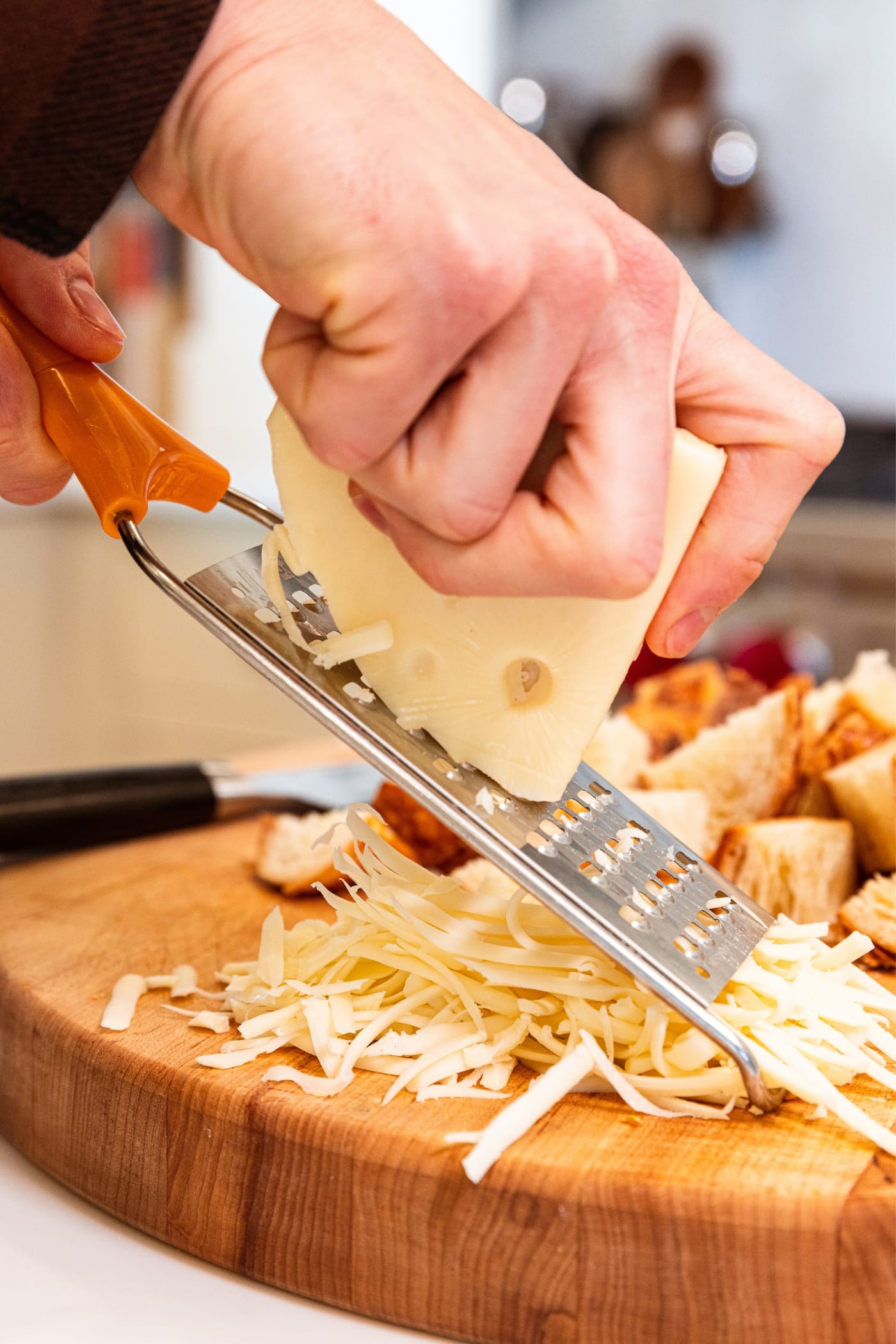 Hand holding block of Swiss cheese shredded on Microplane with pile of already shredded cheese underneath on wood cutting board.