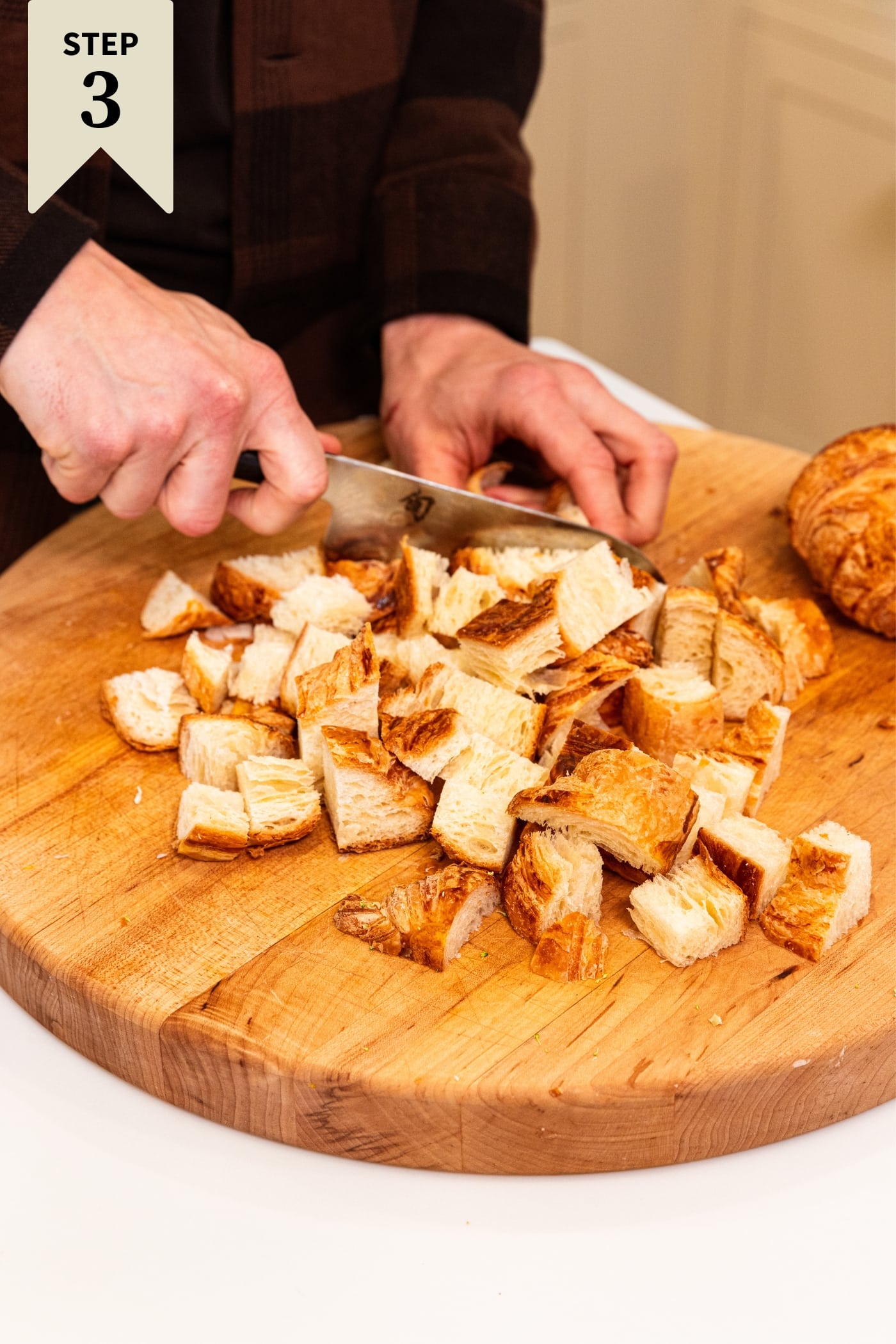 Wood cutting board with hand slicing croissants into small chunks with one croissant in background yet to slice.