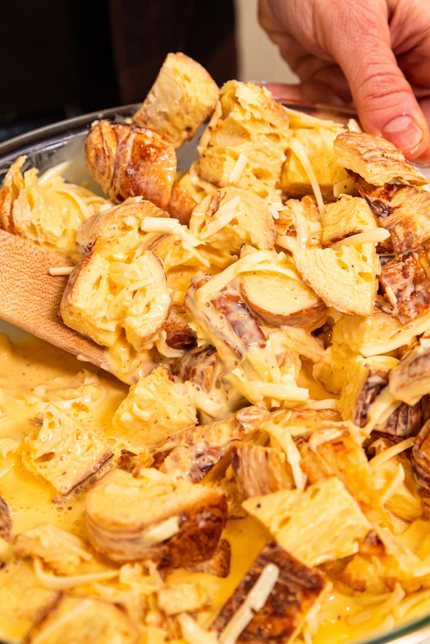 Wooden spatula mixing together cubes of croissant with egg mixture and shredded cheese to form a mixture for egg casserole.