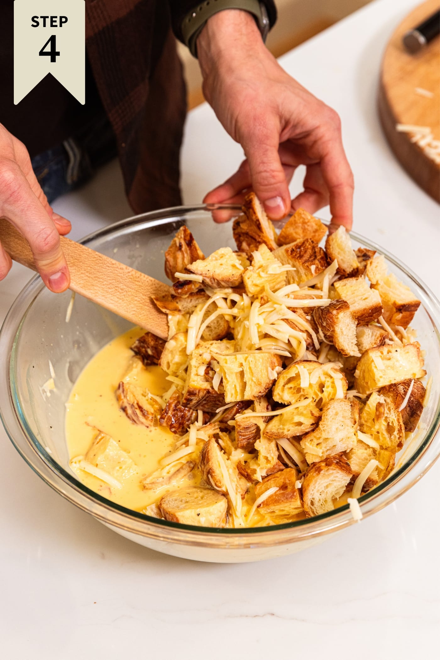 Hand holding wooden spatula mixing together cubed croissants with egg mixture to for a base for egg casserole in glass bowl on white countertop.