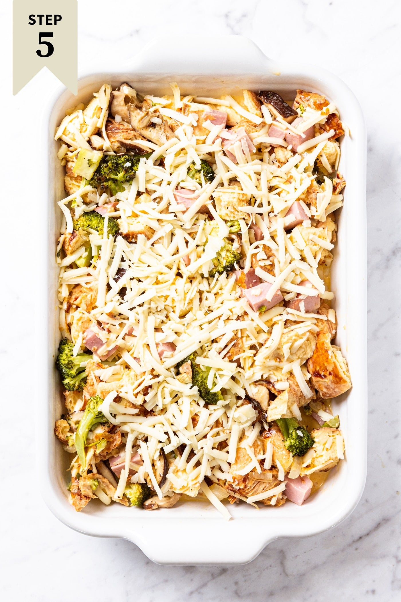 White marble surface with baking dish filled with unbaked egg casserole with shredded cheese on top with broccoli pieces peeking through right before going into the oven.