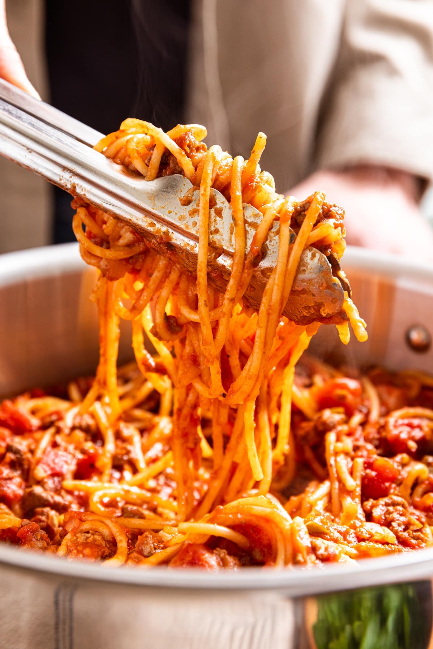 Large metal stockpot with hand holding metal tongs with serving of spaghetti with meat sauce clamped with rest of noodles below.