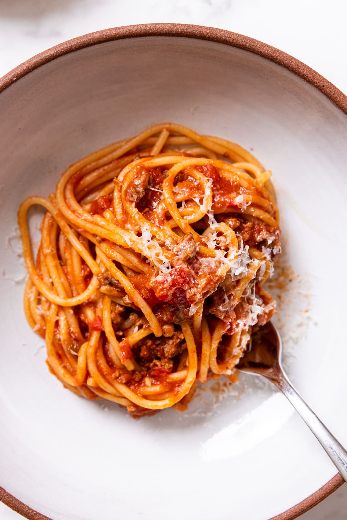 White ceramic bowl filled with swirl of meat sauce with spaghetti with sprinkling of Parmesan cheese on top with fork sticking into the noodles.
