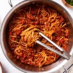 Large metal stockpot filled with meat sauce with spaghetti with tongs sitting on top with swirl of noodles all on marble surface with towel, green herbs, and Parmesan cheese.