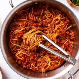 Large metal stockpot filled with meat sauce with spaghetti with tongs sitting on top with swirl of noodles all on marble surface with towel, green herbs, and Parmesan cheese.