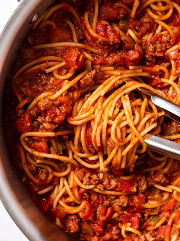 Large metal stockpot filled with spaghetti with meat sauce and tongs resting on top of noodles with swirl of spaghetti all on marble surface.