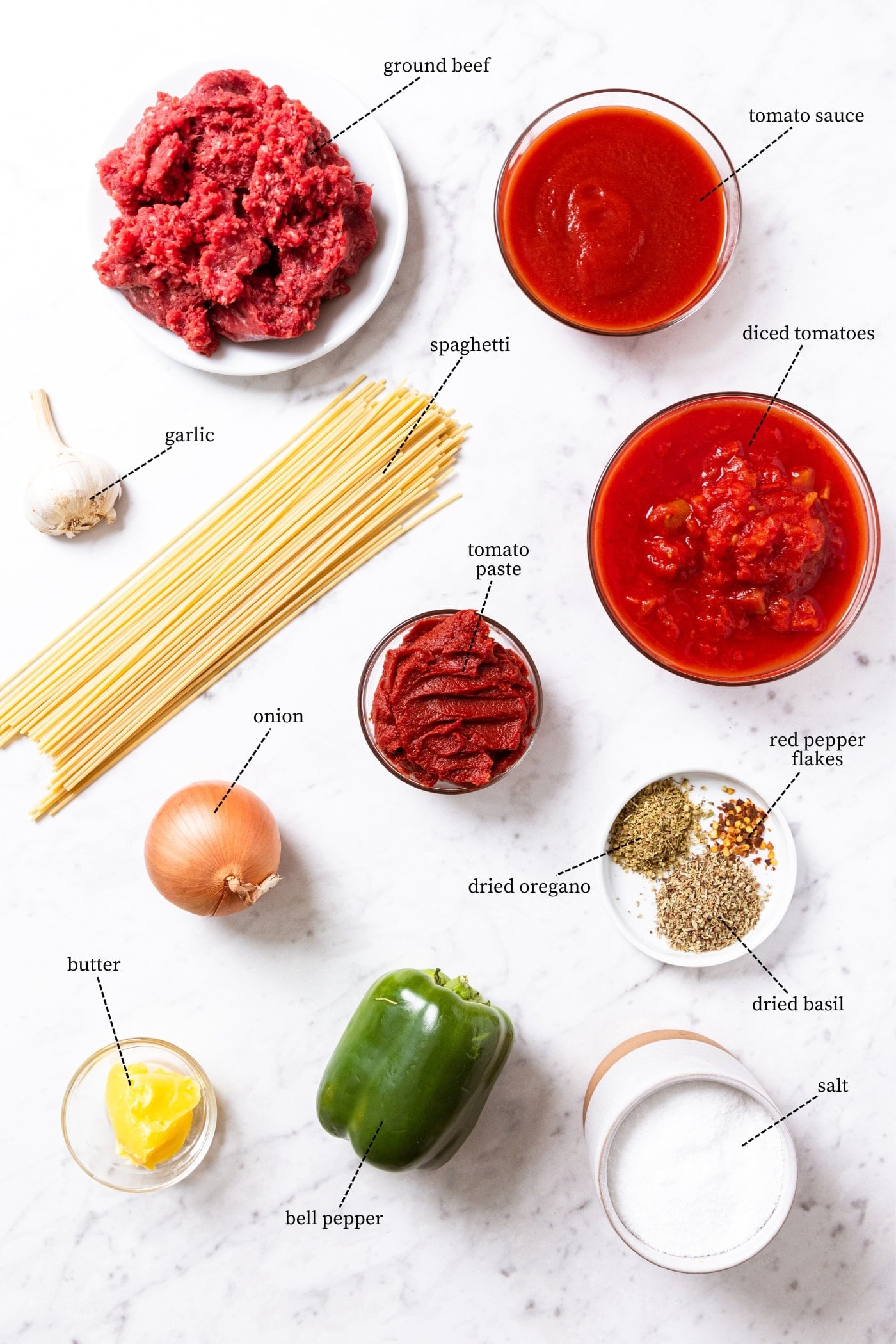 White marble surface with all ingredients needed to make meat sauce with spaghetti including tomato sauce, beef, pasta, green pepper, spices, onion, butter, and more.
